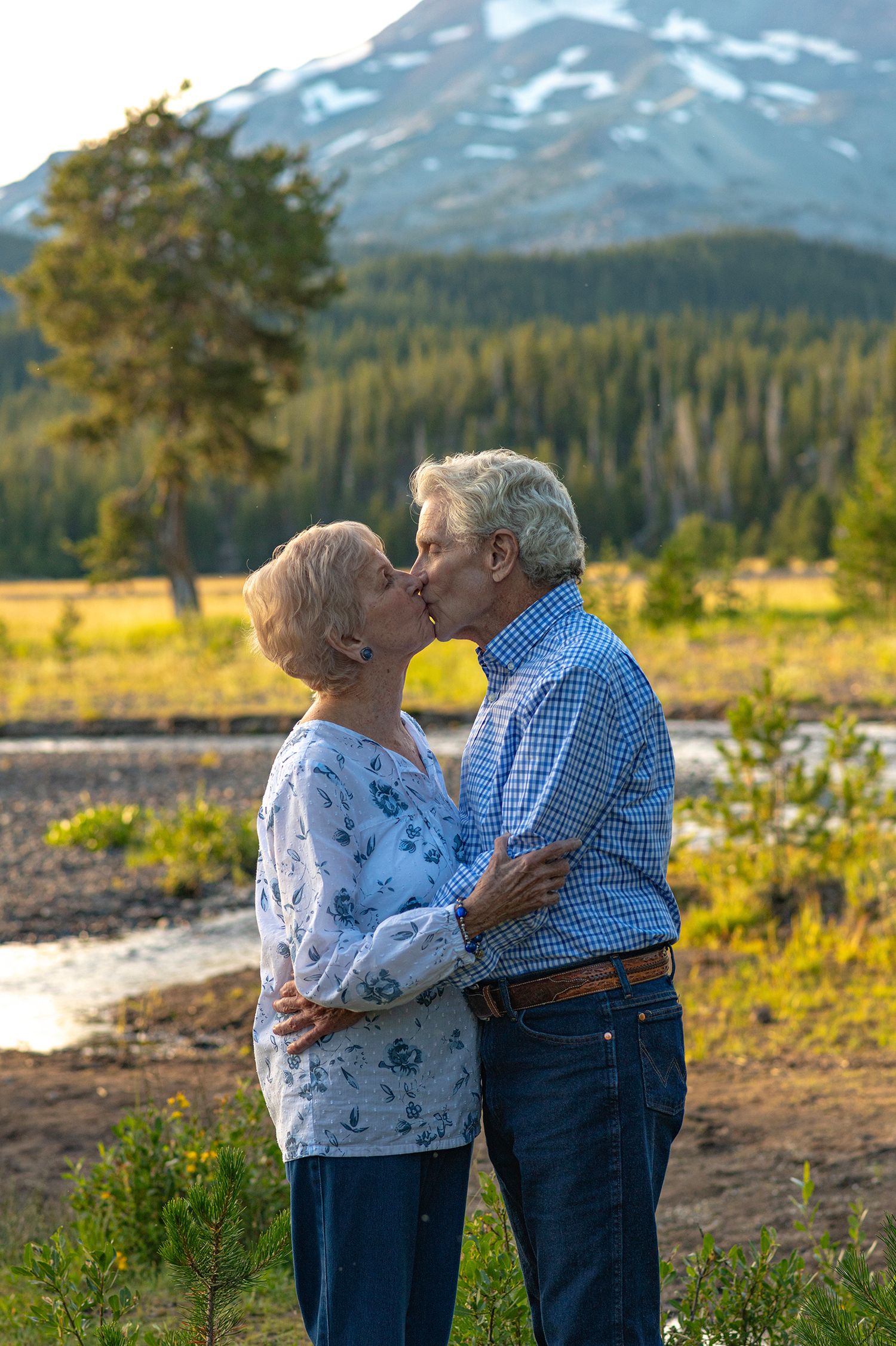 A couple shares a tender kiss in a scenic mountain meadow with snow-capped peaks and evergreen forests in the background.