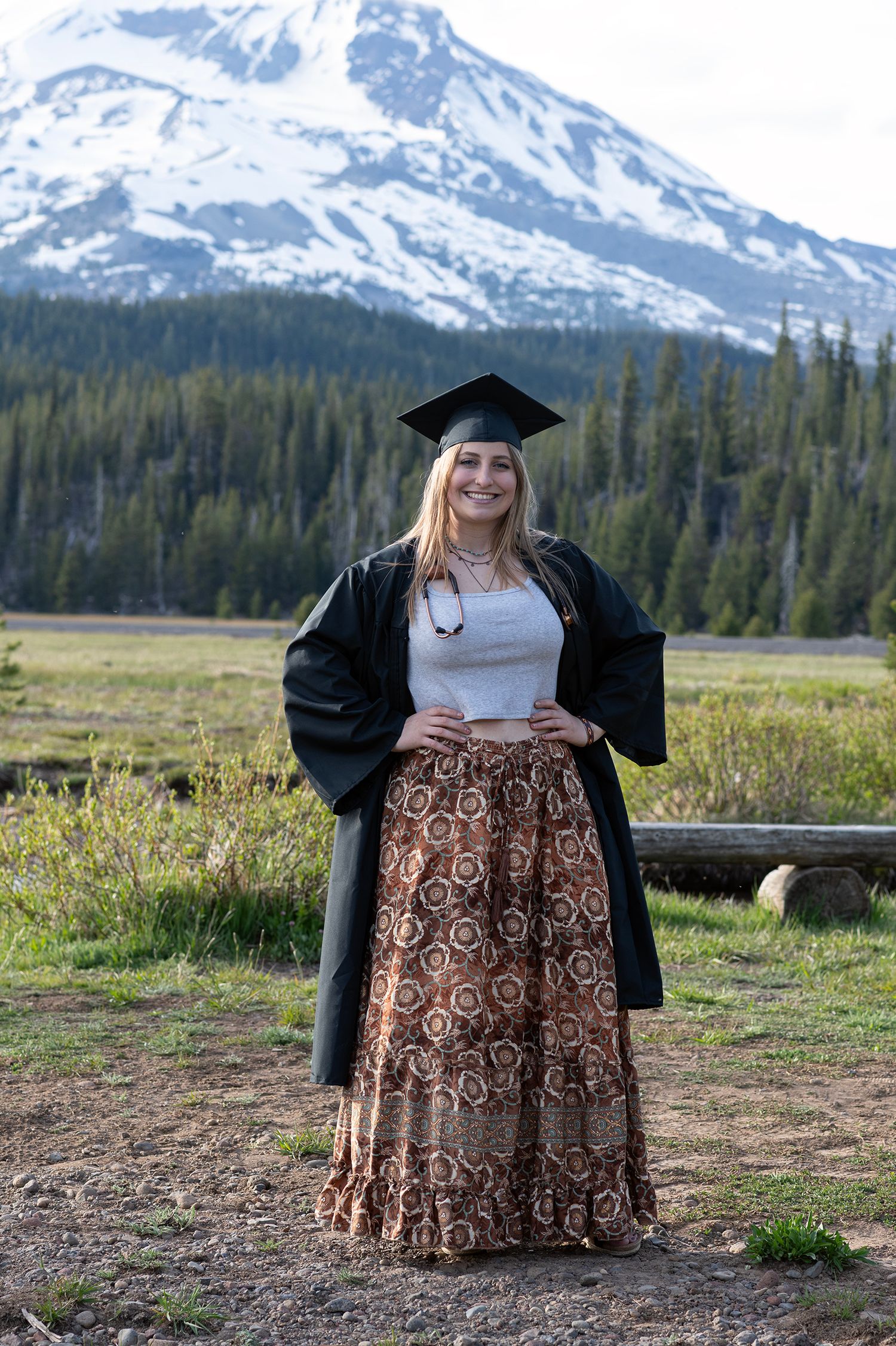 Smiling graduate in cap and gown poses outdoors with snow-capped mountain and pine trees in background.