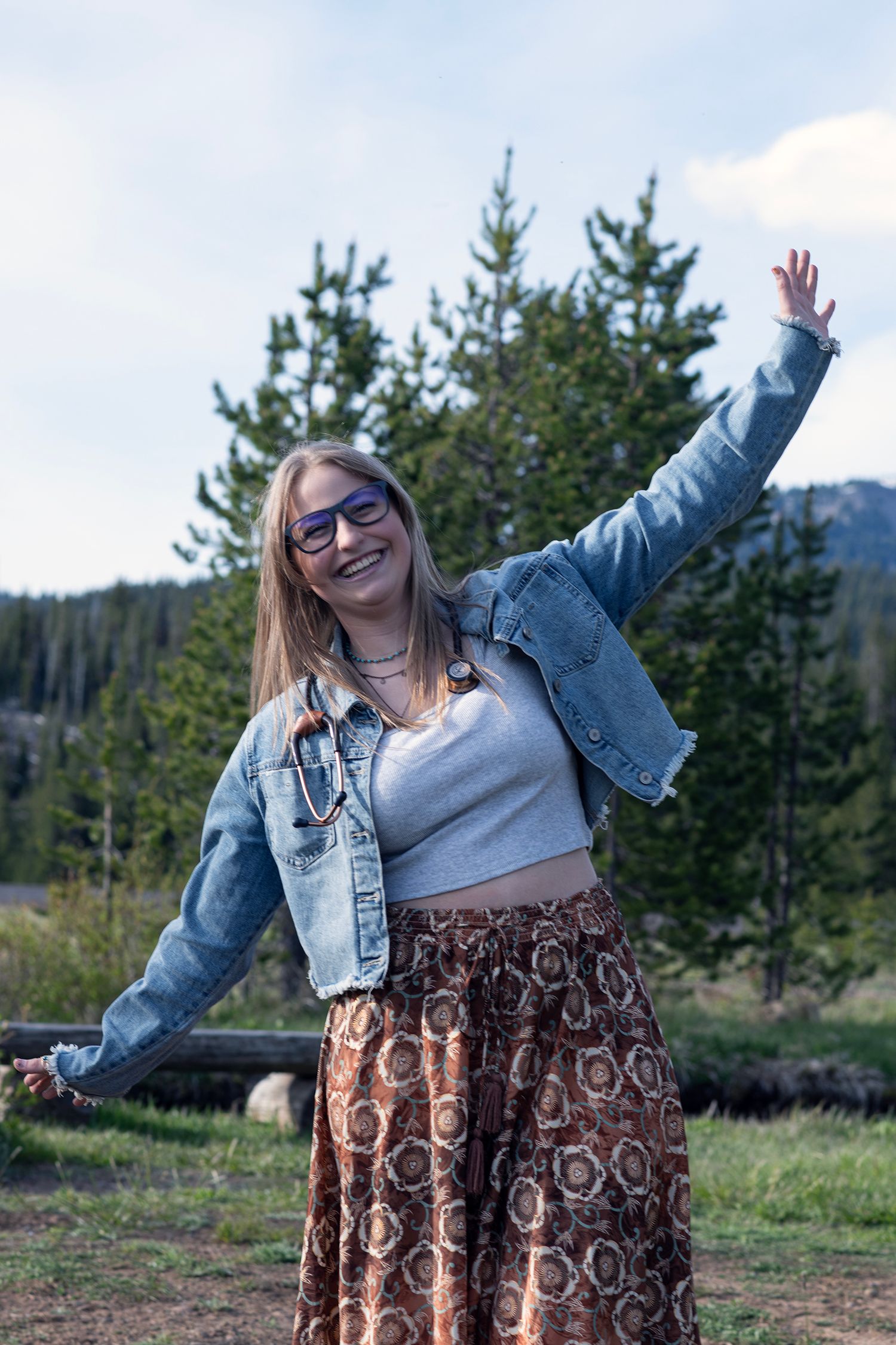 Smiling young woman in denim jacket and boho skirt posing outdoors with arm raised in mountain forest setting.