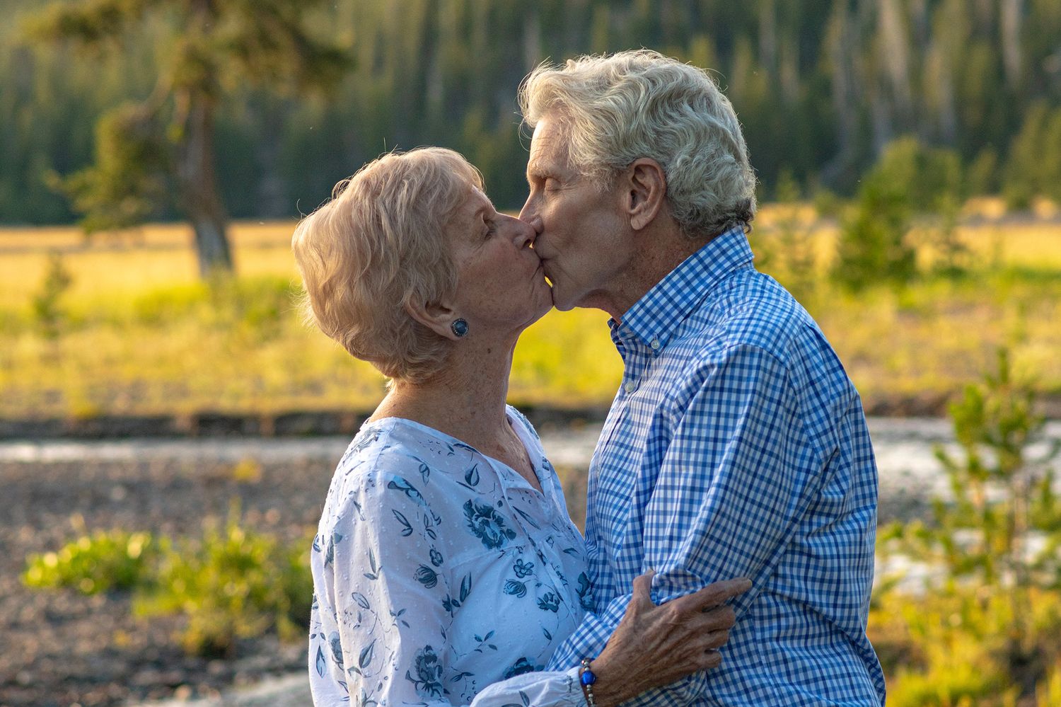 Senior couple sharing a romantic kiss outdoors in a scenic mountain meadow with wildflowers at sunset.