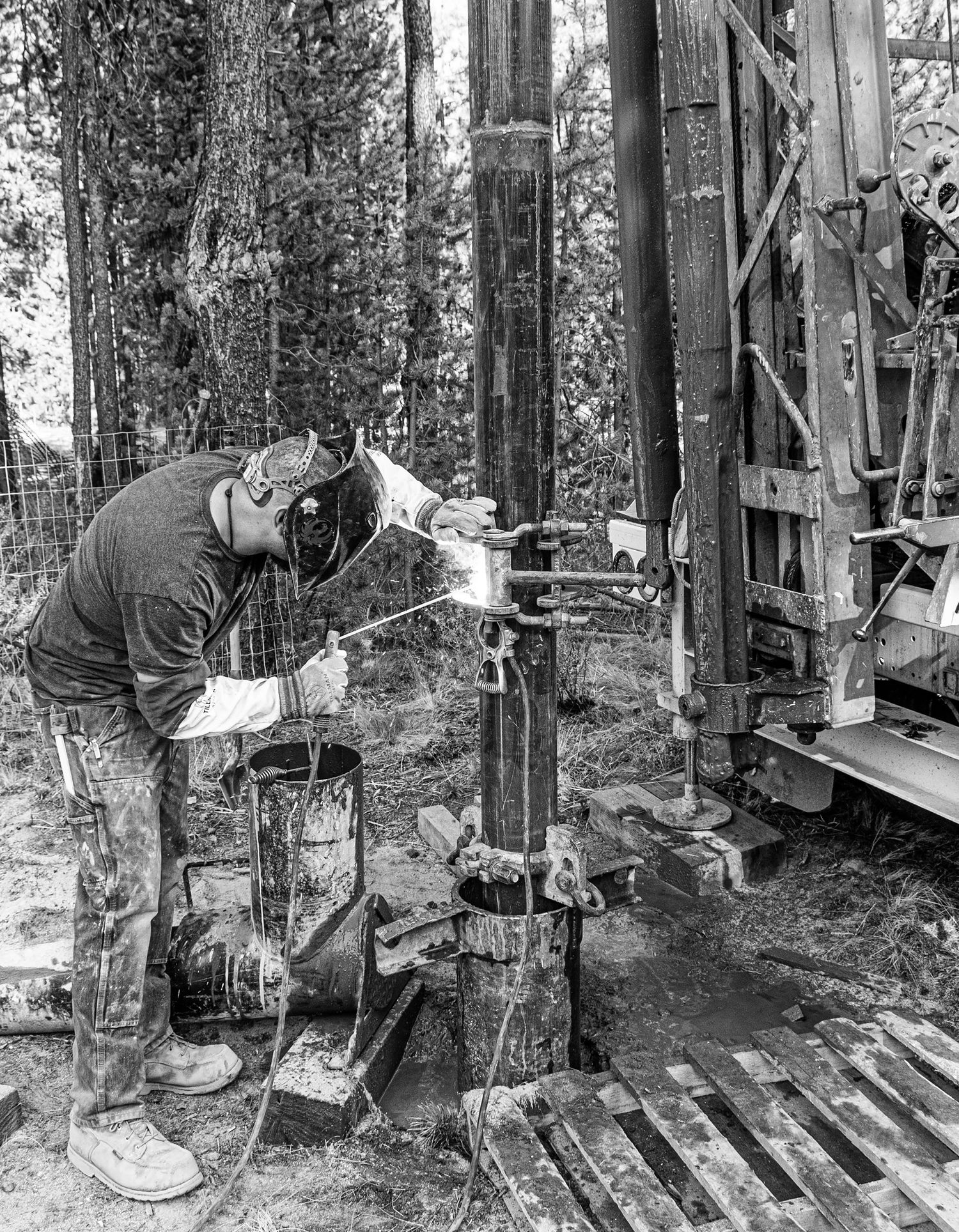 Worker operating drilling equipment in a forest setting with protective gear.