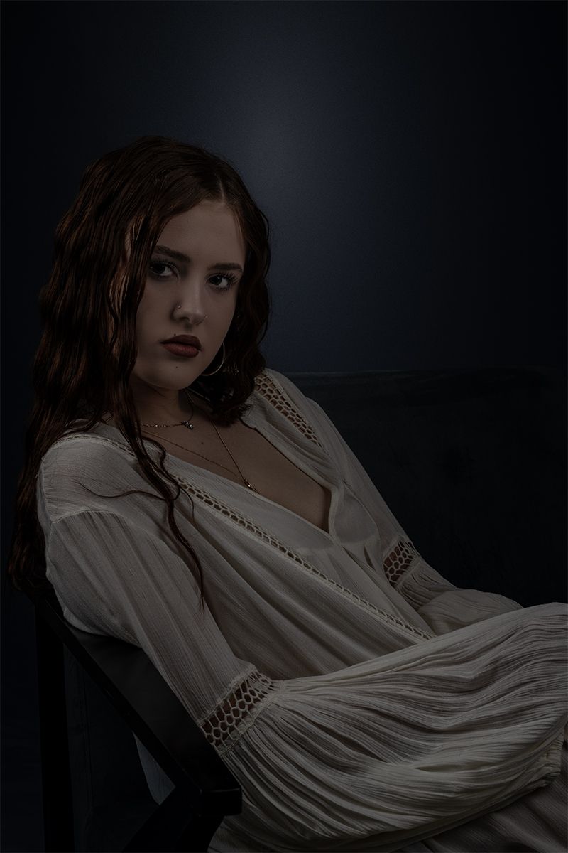 A moody fashion portrait shows a figure in a white flowing dress reclining against a dark background.