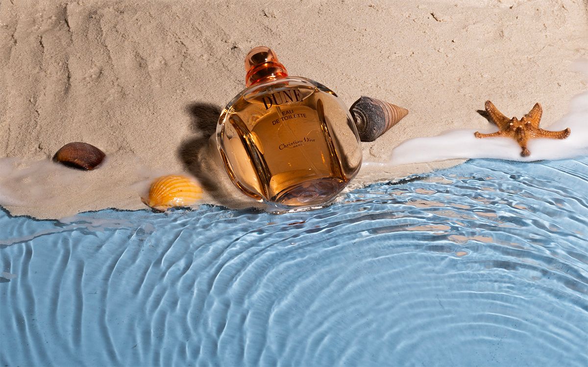 Bottle of perfume arranged artistically on sand with seashells and blue water.