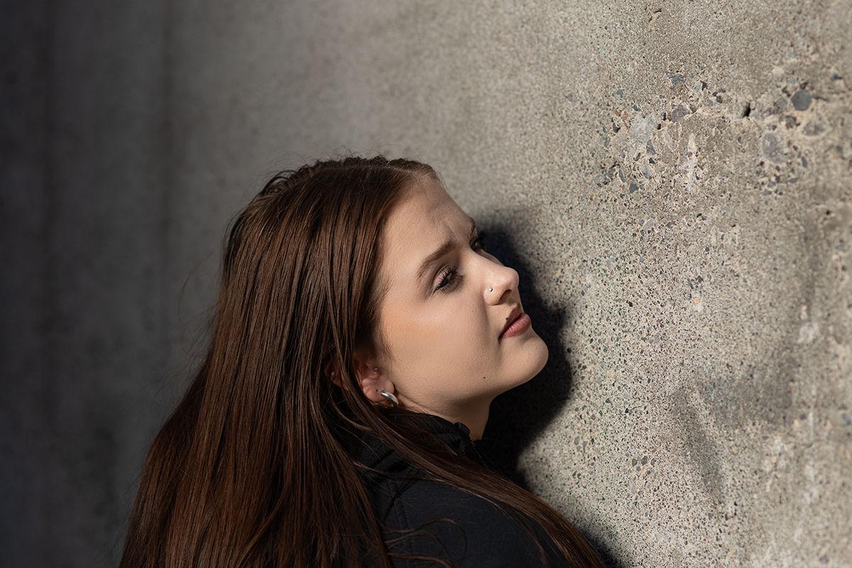 A person with long brown hair leans against a textured concrete wall in dramatic lighting.