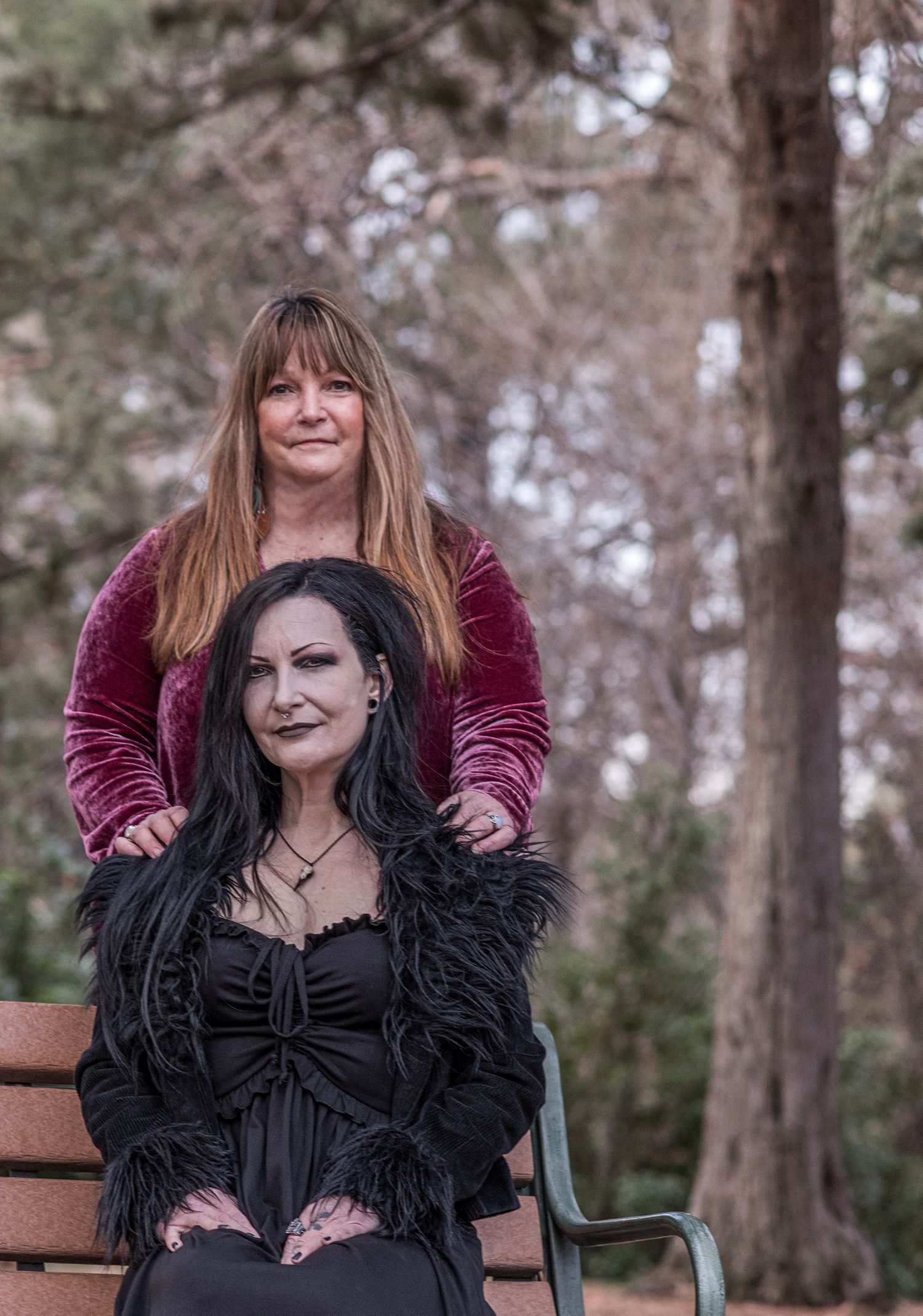 Two people in contrasting black and burgundy velvet outfits pose together in a wooded outdoor setting.