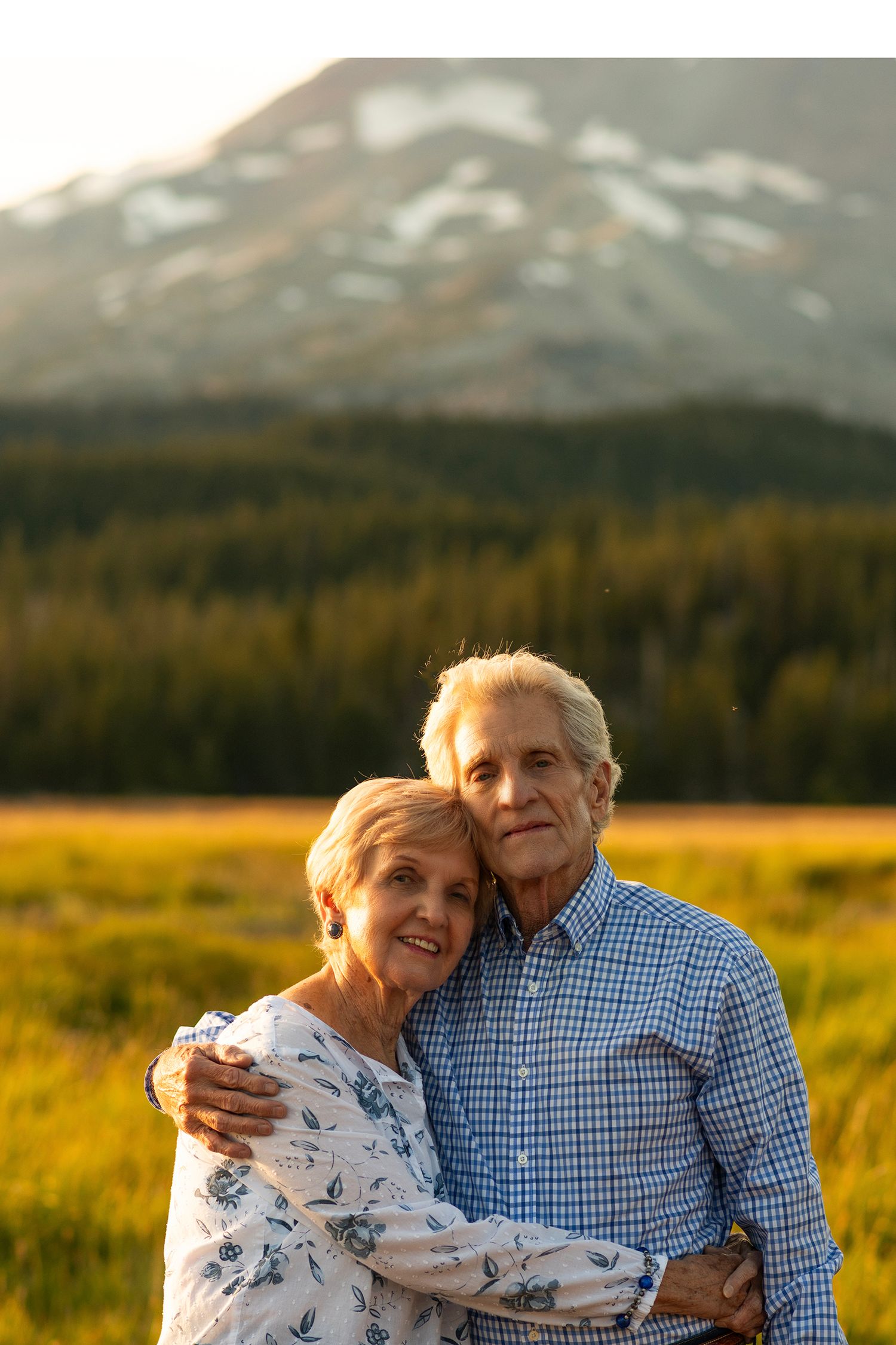 An elderly couple embraces while standing in a golden meadow with snow-capped mountains in the background at sunset.
