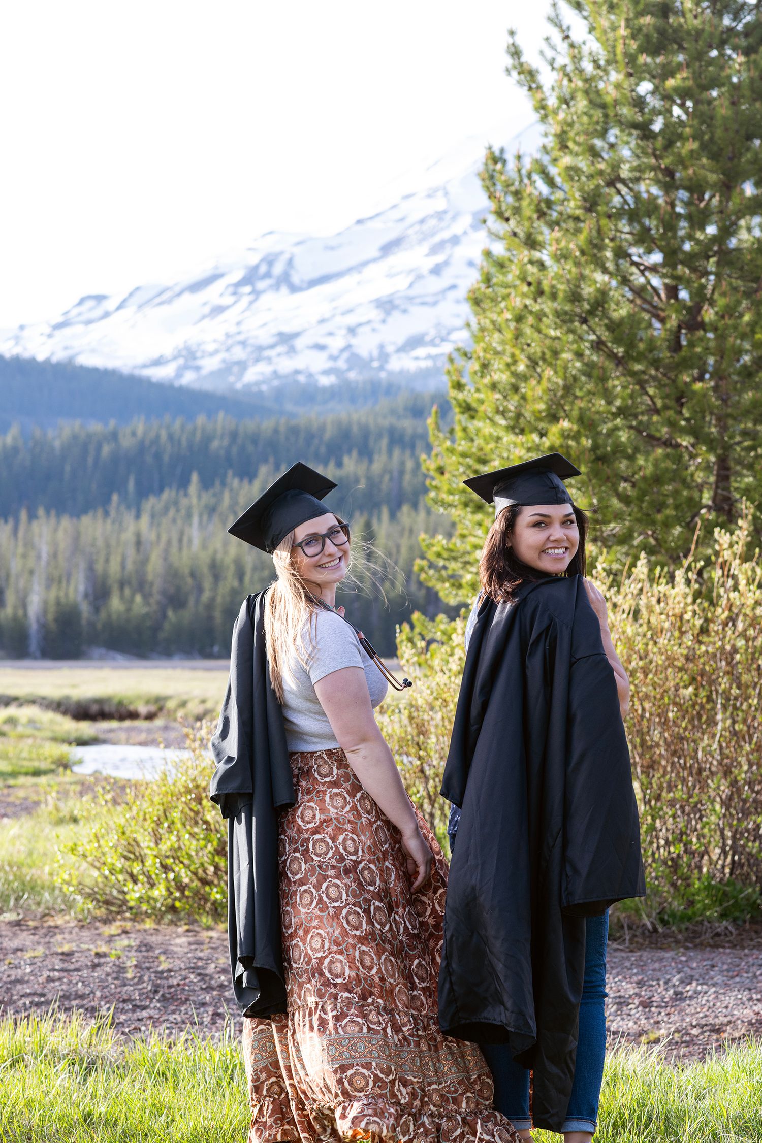 Two women in graduation caps and gowns pose outdoors with a snowy mountain and pine trees in the background.