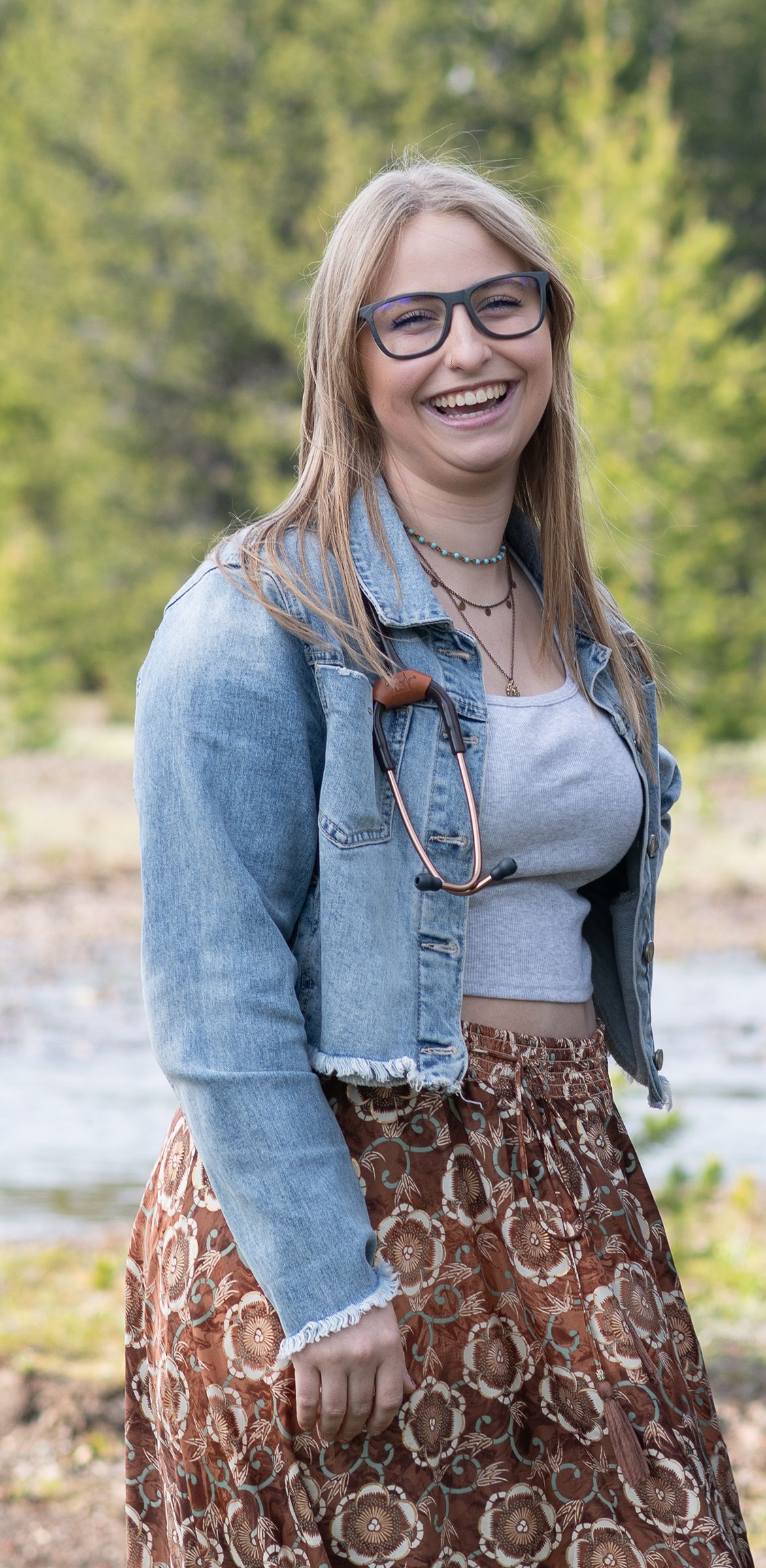 Smiling young blonde woman wearing glasses, denim jacket, and floral skirt posing outdoors near trees.