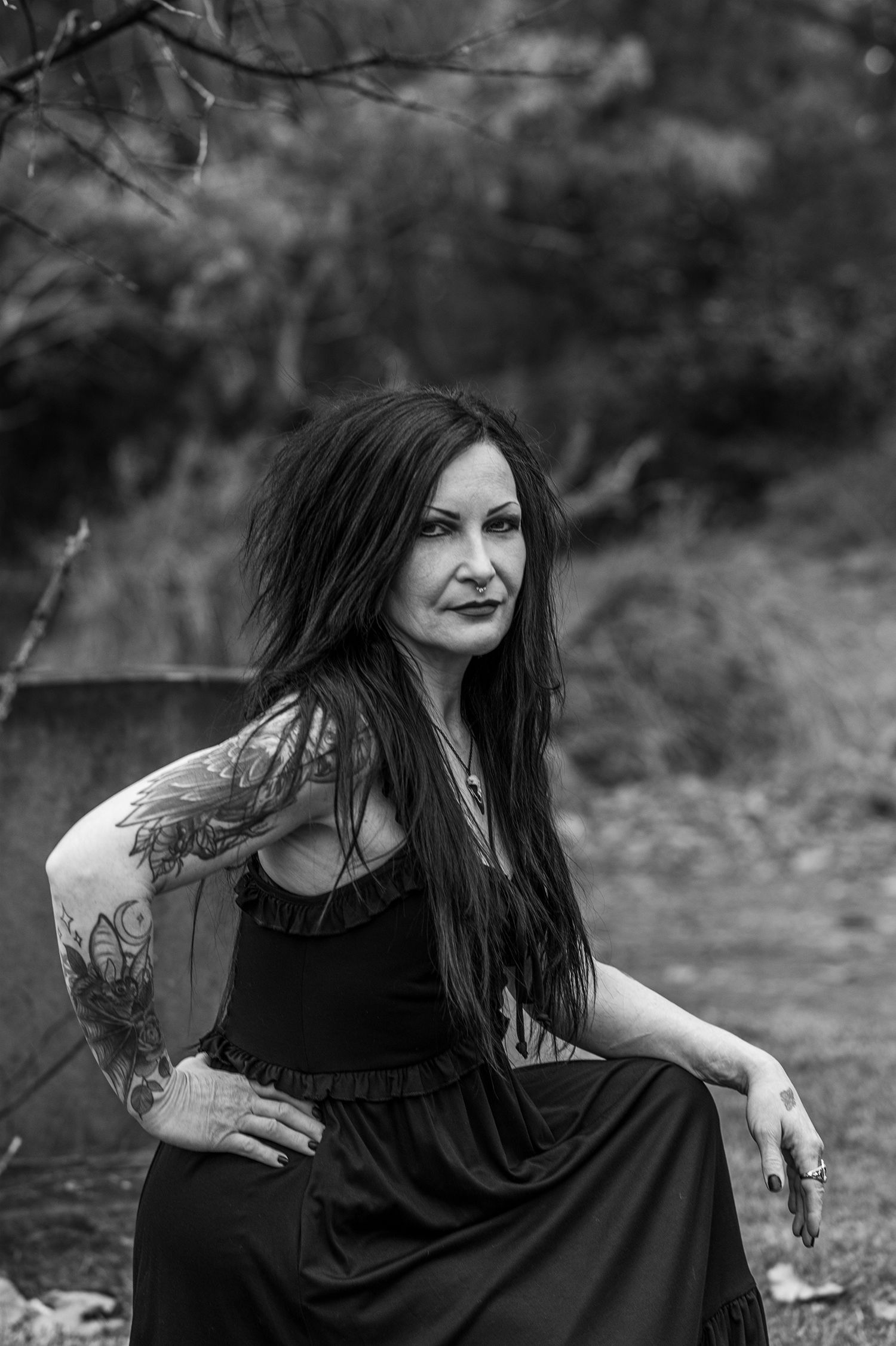 Black and white portrait of a woman in a dark dress with tattoos sitting outdoors in a natural setting.