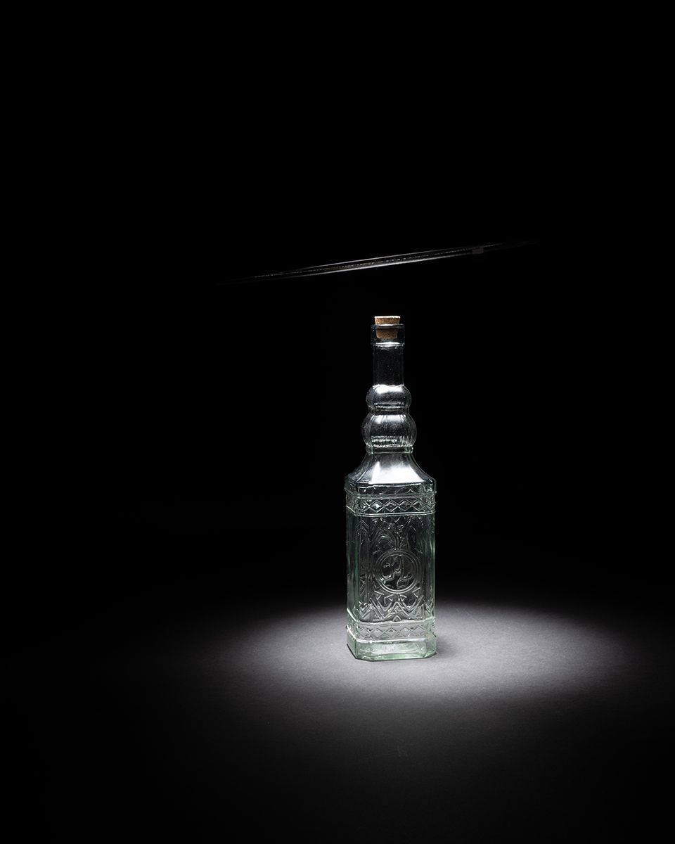 A glass bottle illuminated by dramatic spotlight fades into darkness across three sequential artistic photographs.