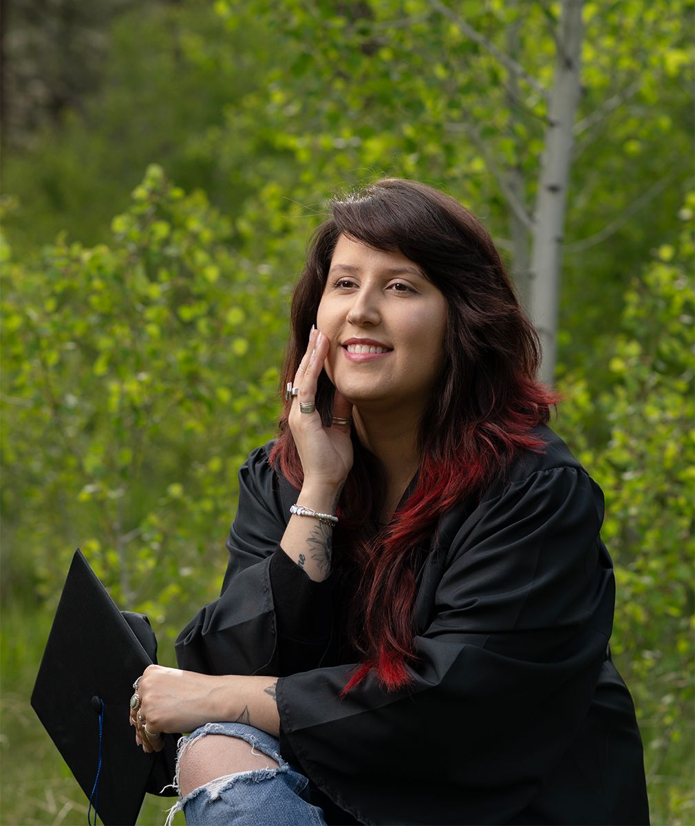 Graduate in black gown with colorful hair poses outdoors against green foliage.