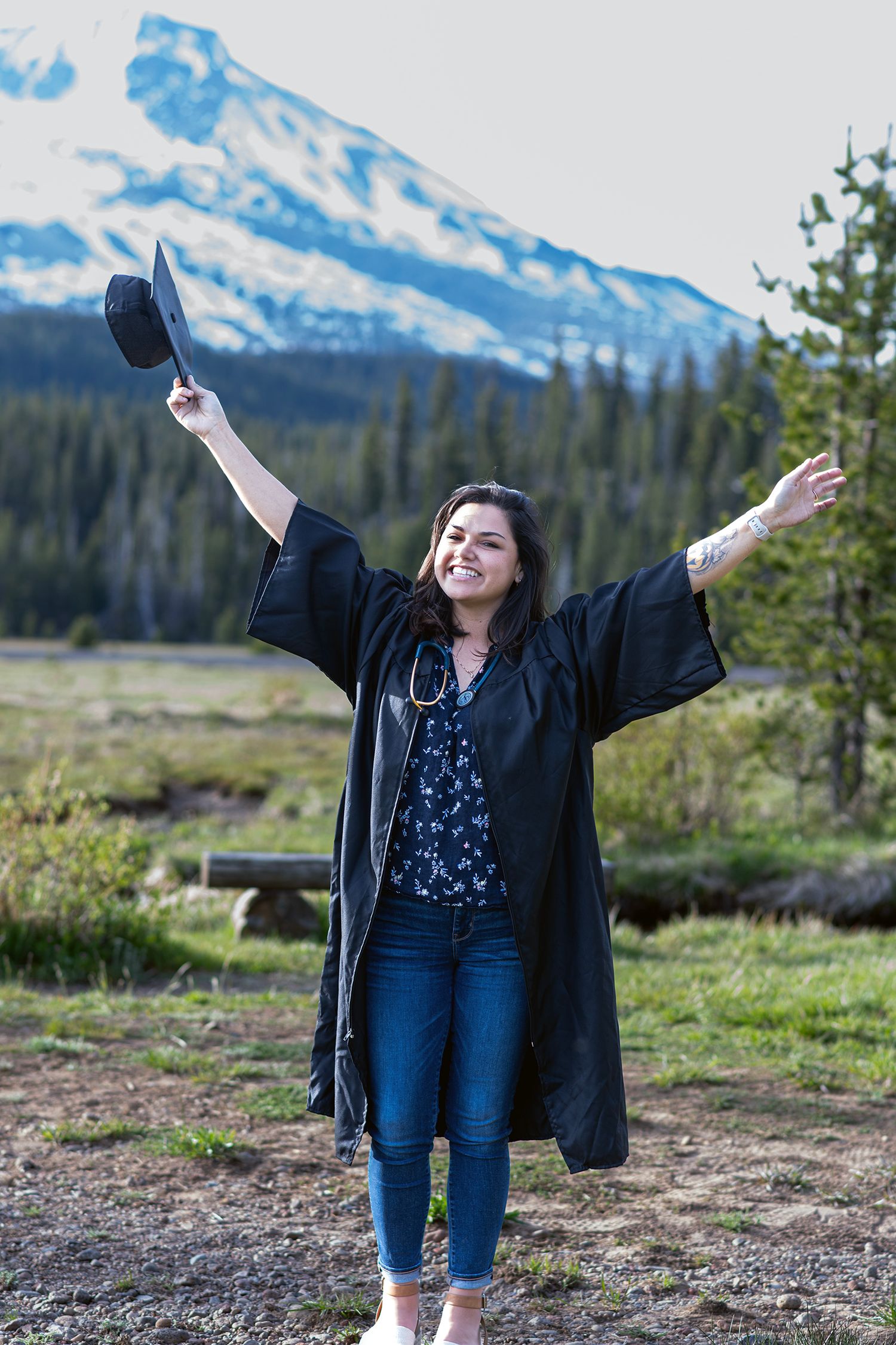 Joyful graduate in black gown raises arms triumphantly outdoors with snow-capped mountain backdrop.