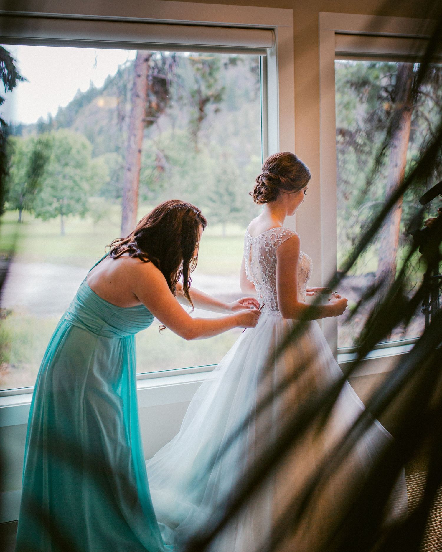 A bridesmaid in turquoise helps adjust a wedding dress by a large window.