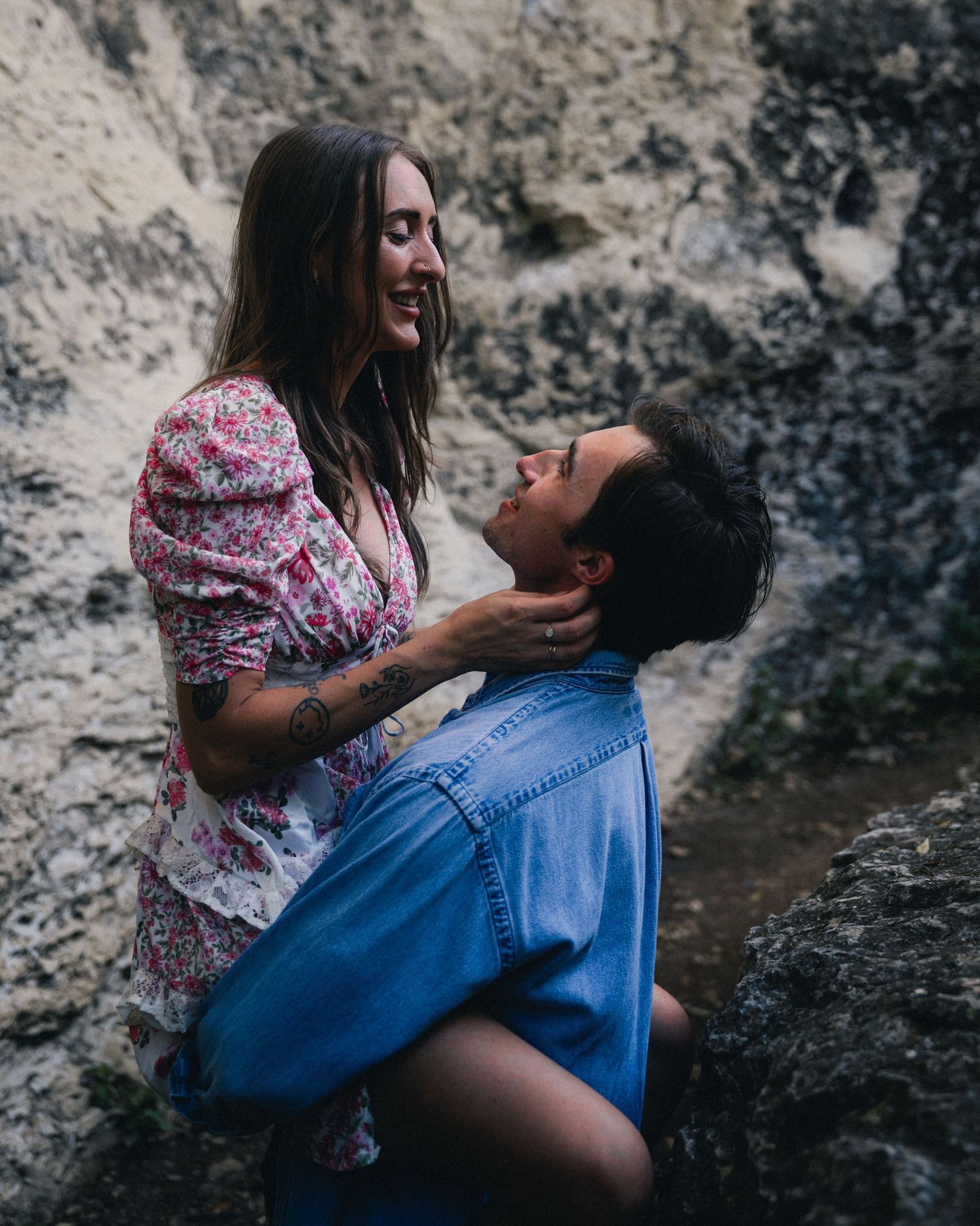 A couple in romantic embrace near rocky cliffs, with one wearing a floral dress and the other in denim.