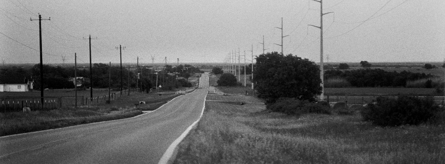 A black and white photograph shows a long empty rural road stretching into the distance with telephone poles along its sides.