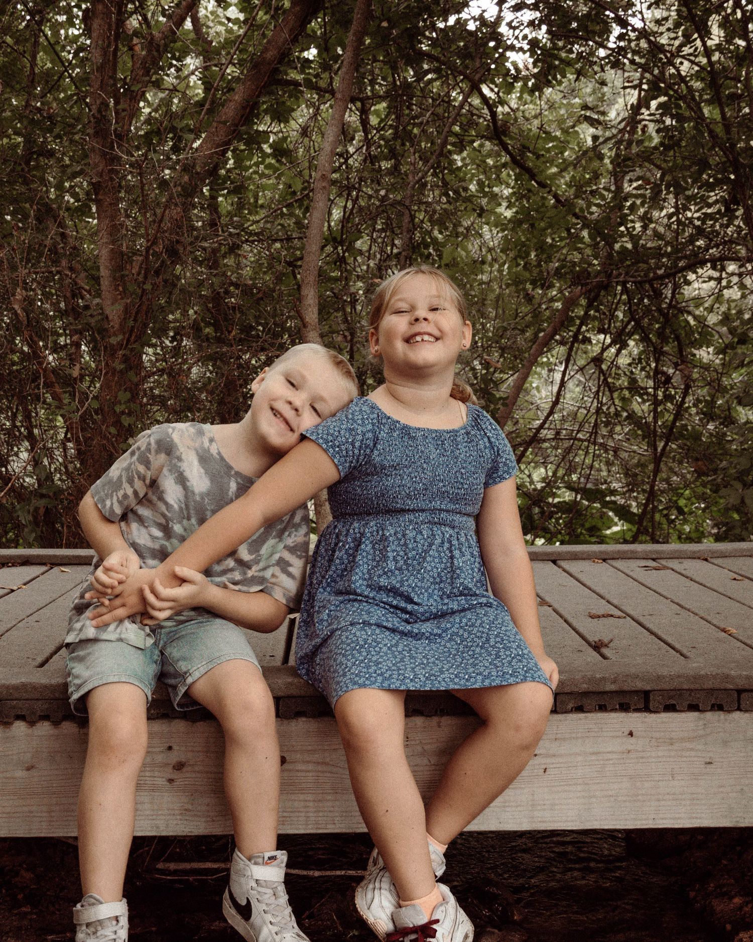 Two children share a playful moment while sitting on a wooden bench surrounded by trees.