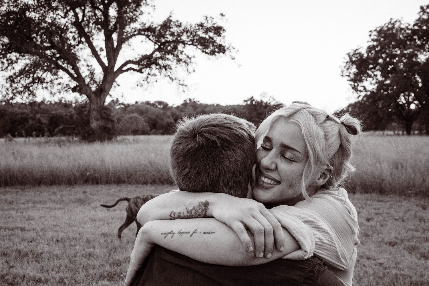 Black and white photo showing an intimate hug between two people outdoors in a grassy field with trees in background.