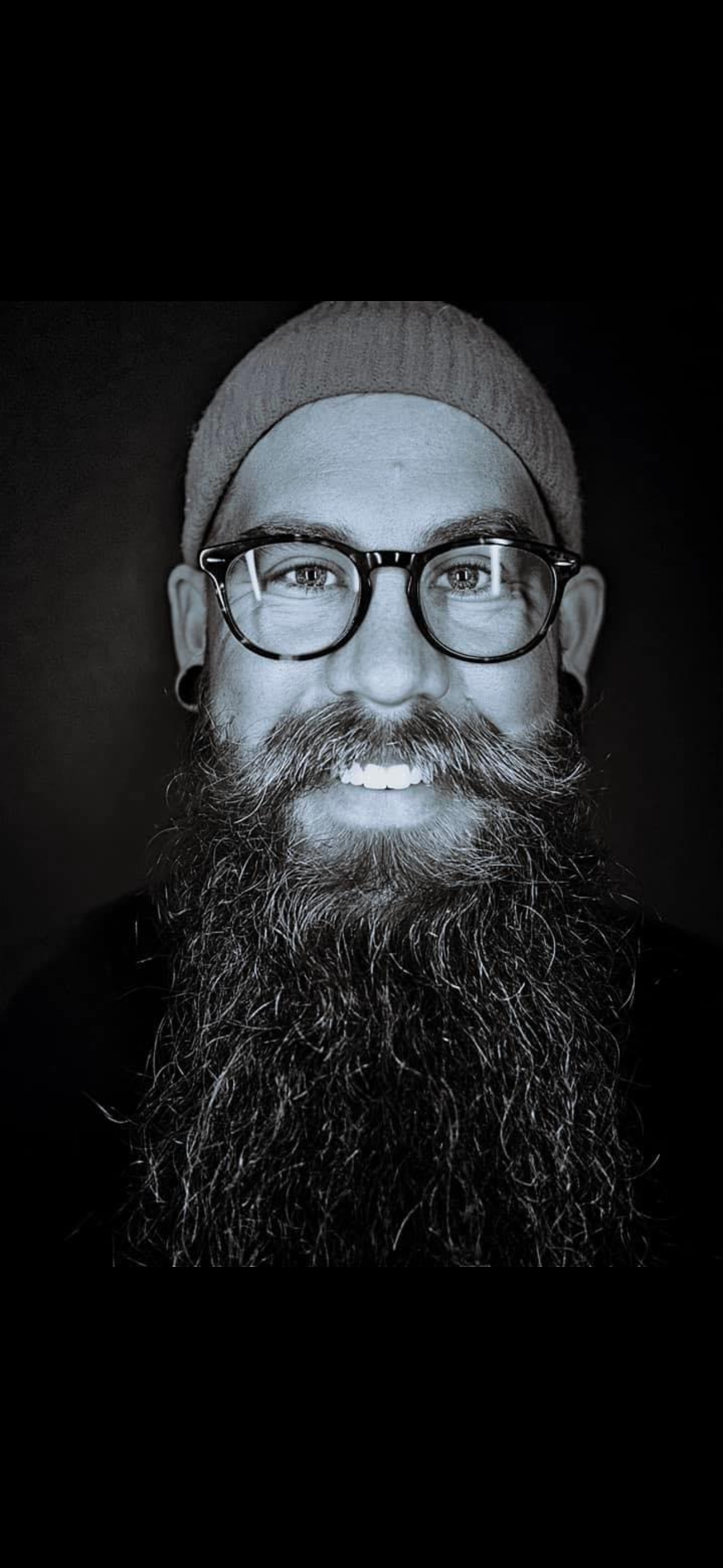 Black and white portrait of Lance wearing glasses and a beanie, featuring a long full beard and bright smile.