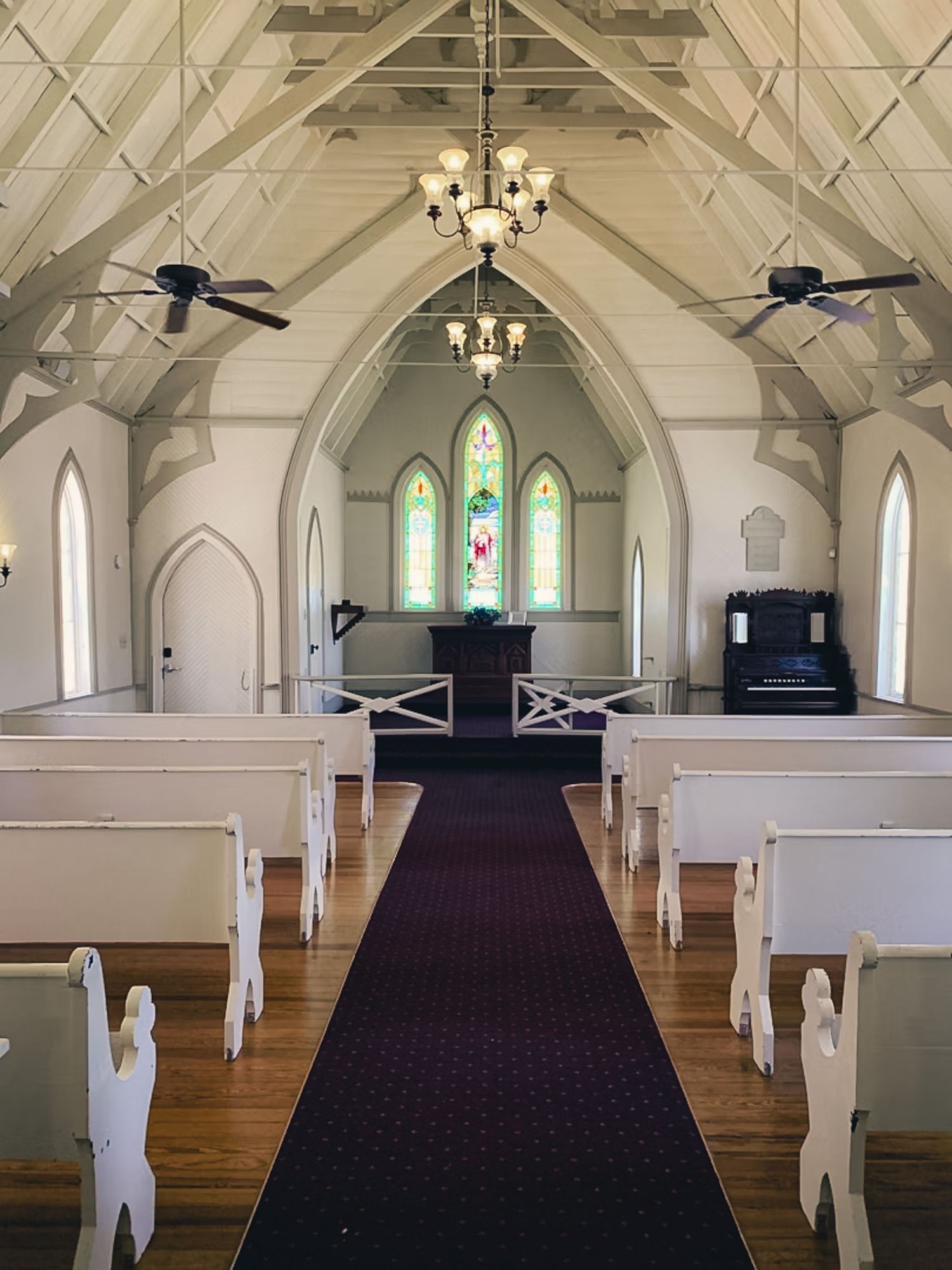 Interior view of a small chapel with white walls, vaulted ceiling, stained glass windows, chandeliers and wooden pews.