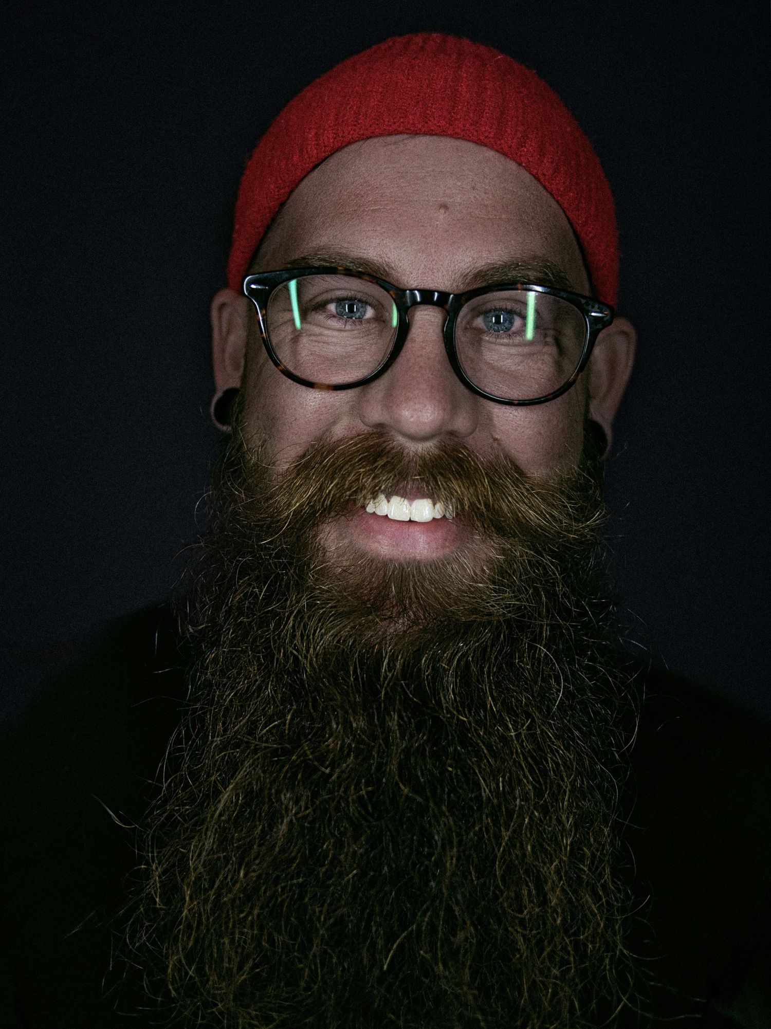 Lance wearing glasses and red beanie hat smiles against dark background in portrait photo.