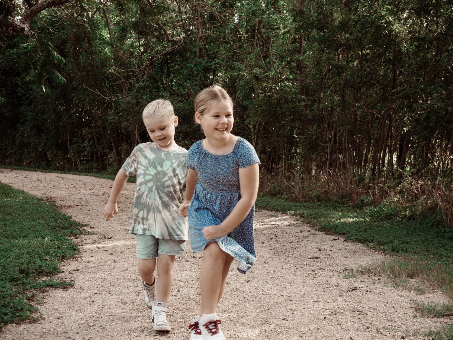 Two children running playfully on a dirt path through the woods.