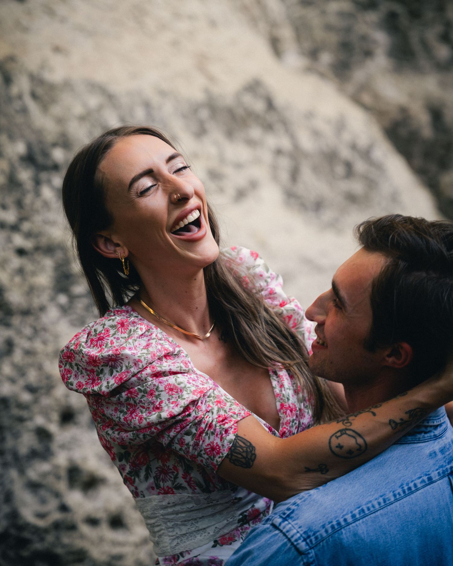 Two people share a joyful intimate moment together against a rocky background while wearing floral and denim.