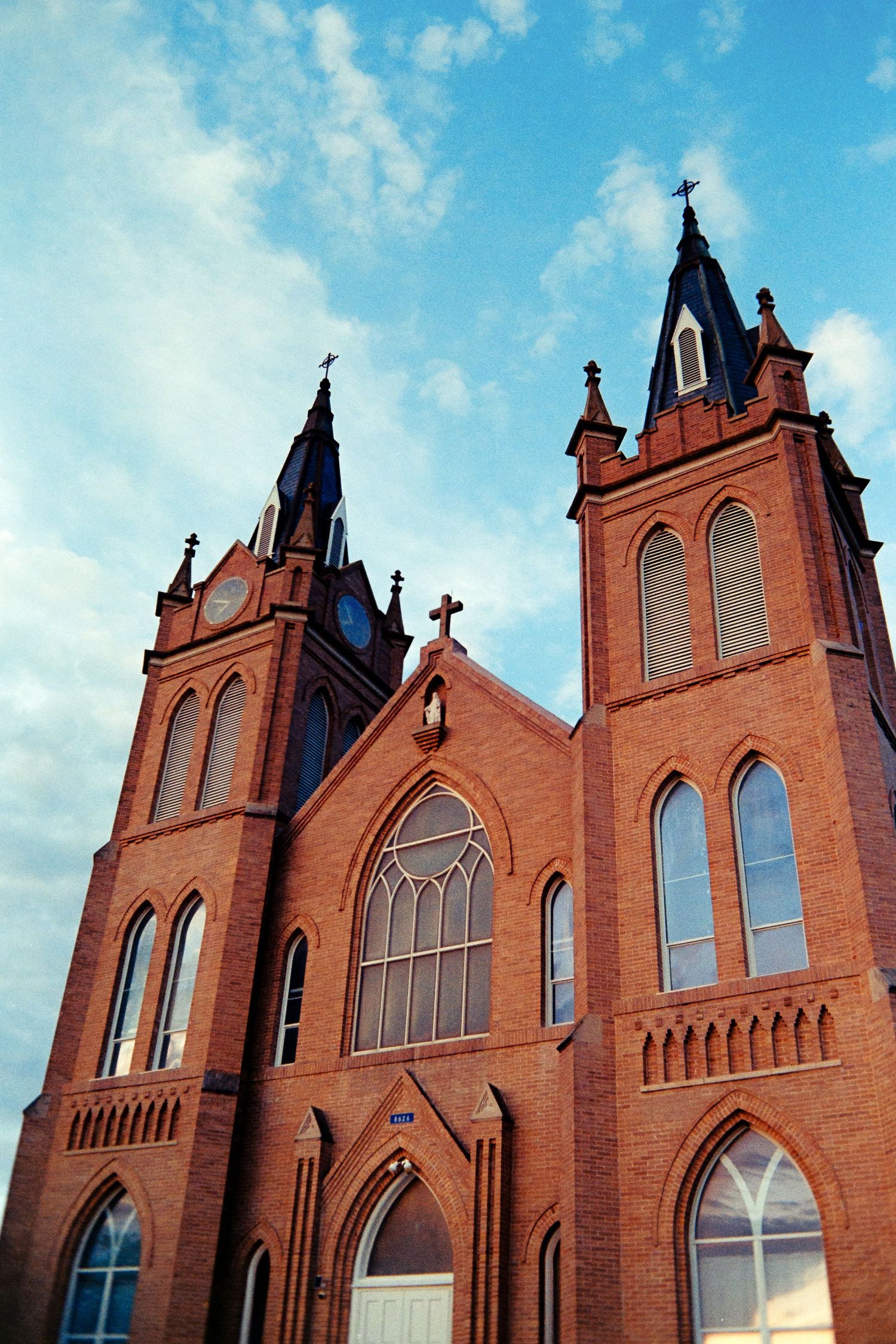 Symmetrical red brick Gothic Revival church with tall twin spires and arched windows against a blue cloudy sky.