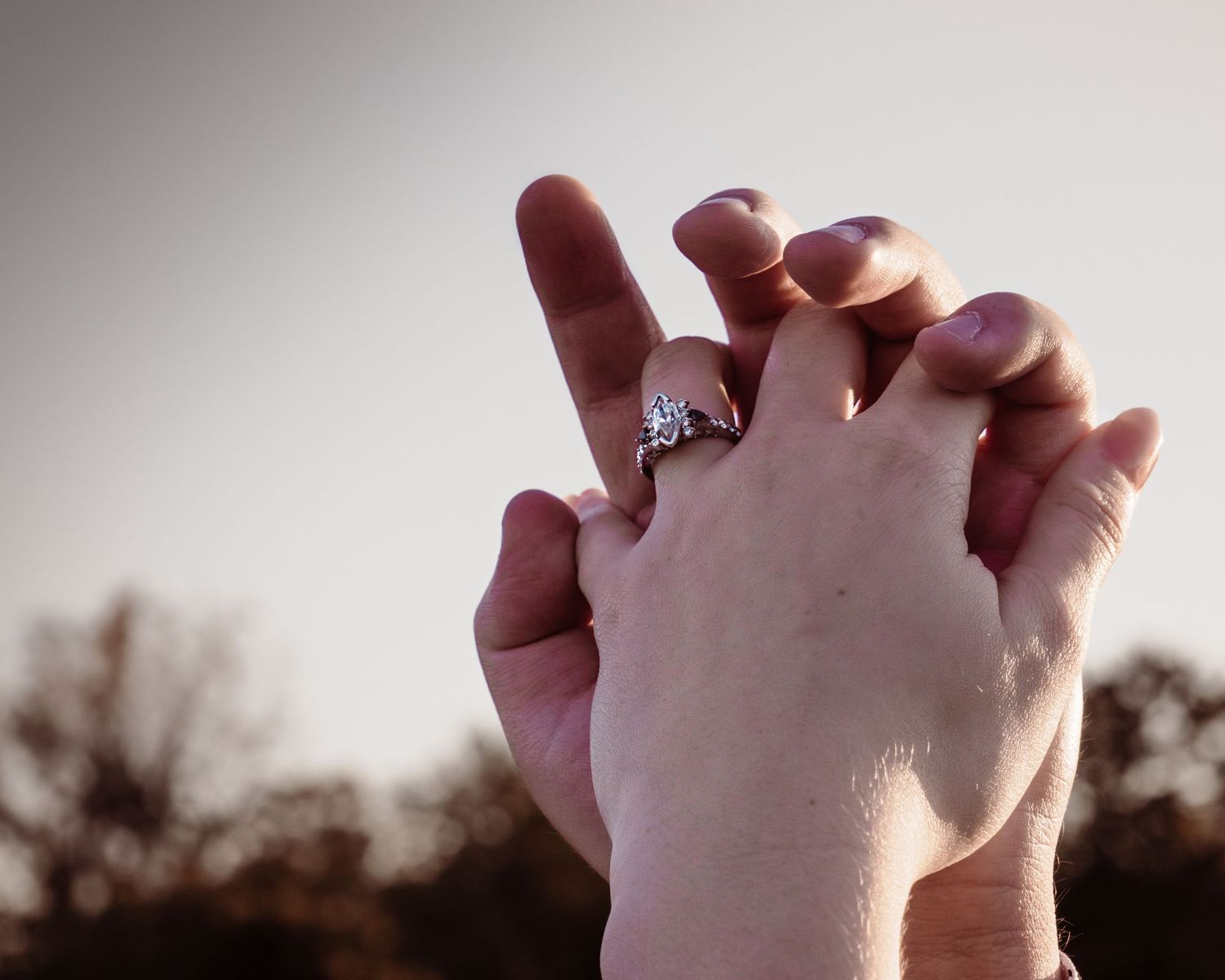 Close-up shot of clasped hands with an engagement ring silhouetted against sunset sky.