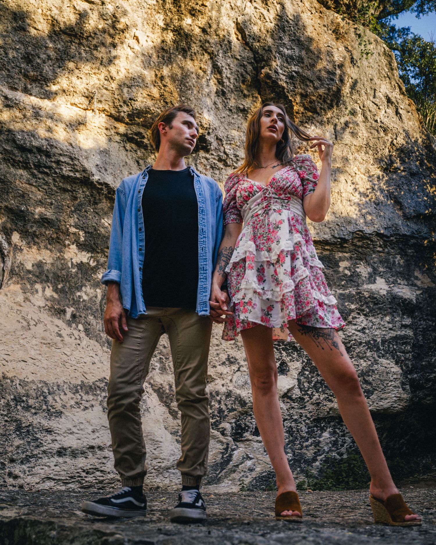 A couple poses casually against a rocky cliff wall during a golden hour photoshoot, combining casual and dressy style.