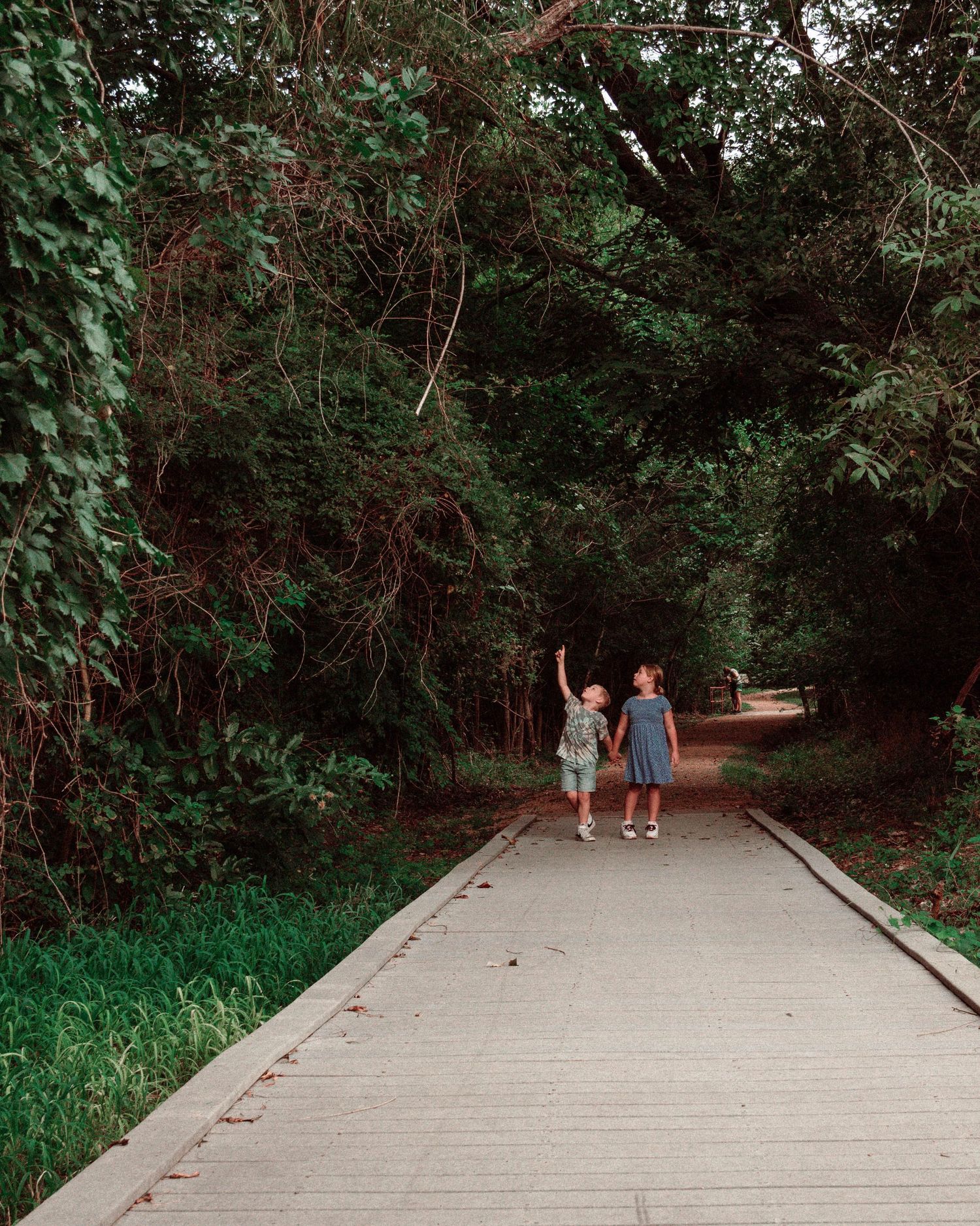 Two children walk together on a paved pathway through a lush green wooded area on a sunny day.