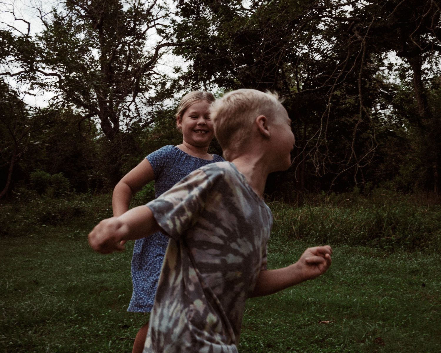 Two children enjoy playing and laughing together on a grassy lawn surrounded by trees at dusk.