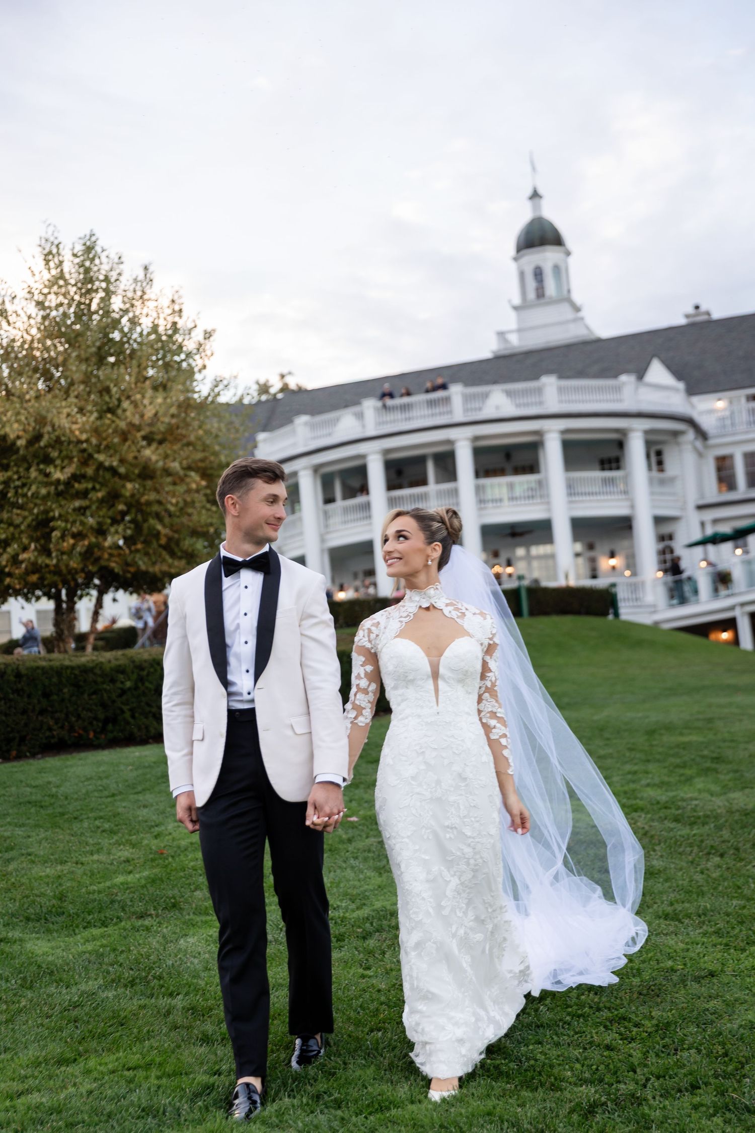 Newlywed couple walks hand in hand on a lush green lawn with a grand white colonial-style building in the background.