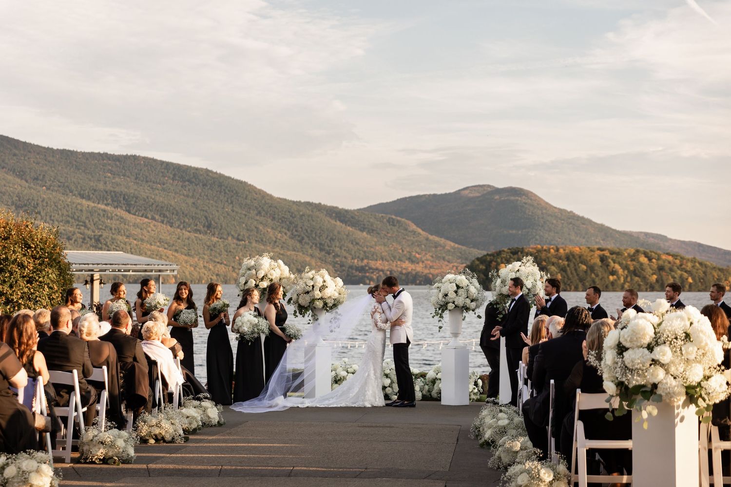 A panoramic view of an outdoor wedding ceremony overlooking a scenic mountain lake with white floral arrangements.