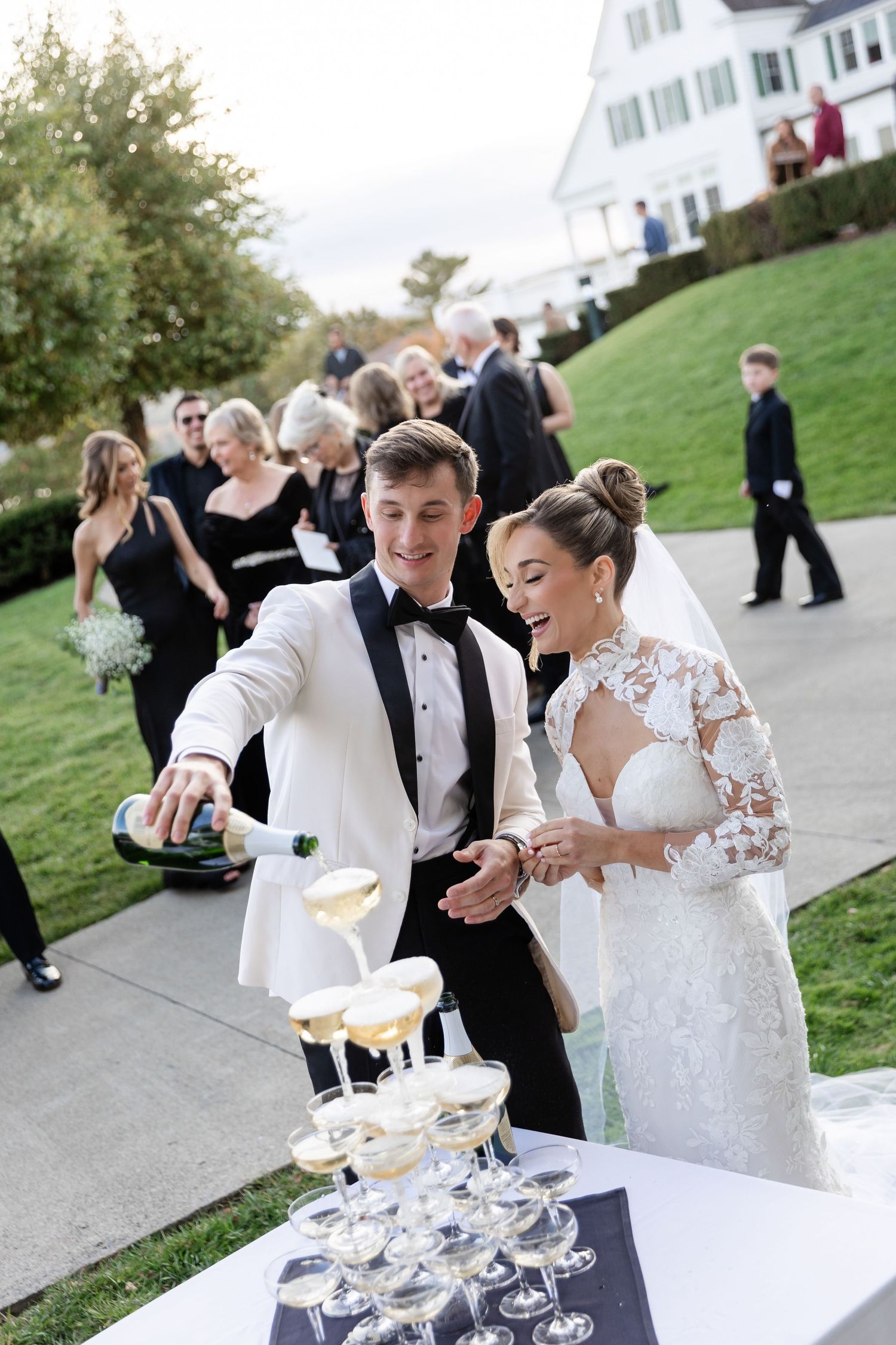Newlywed couple pours champagne into a tower of glasses during their outdoor wedding reception celebration.