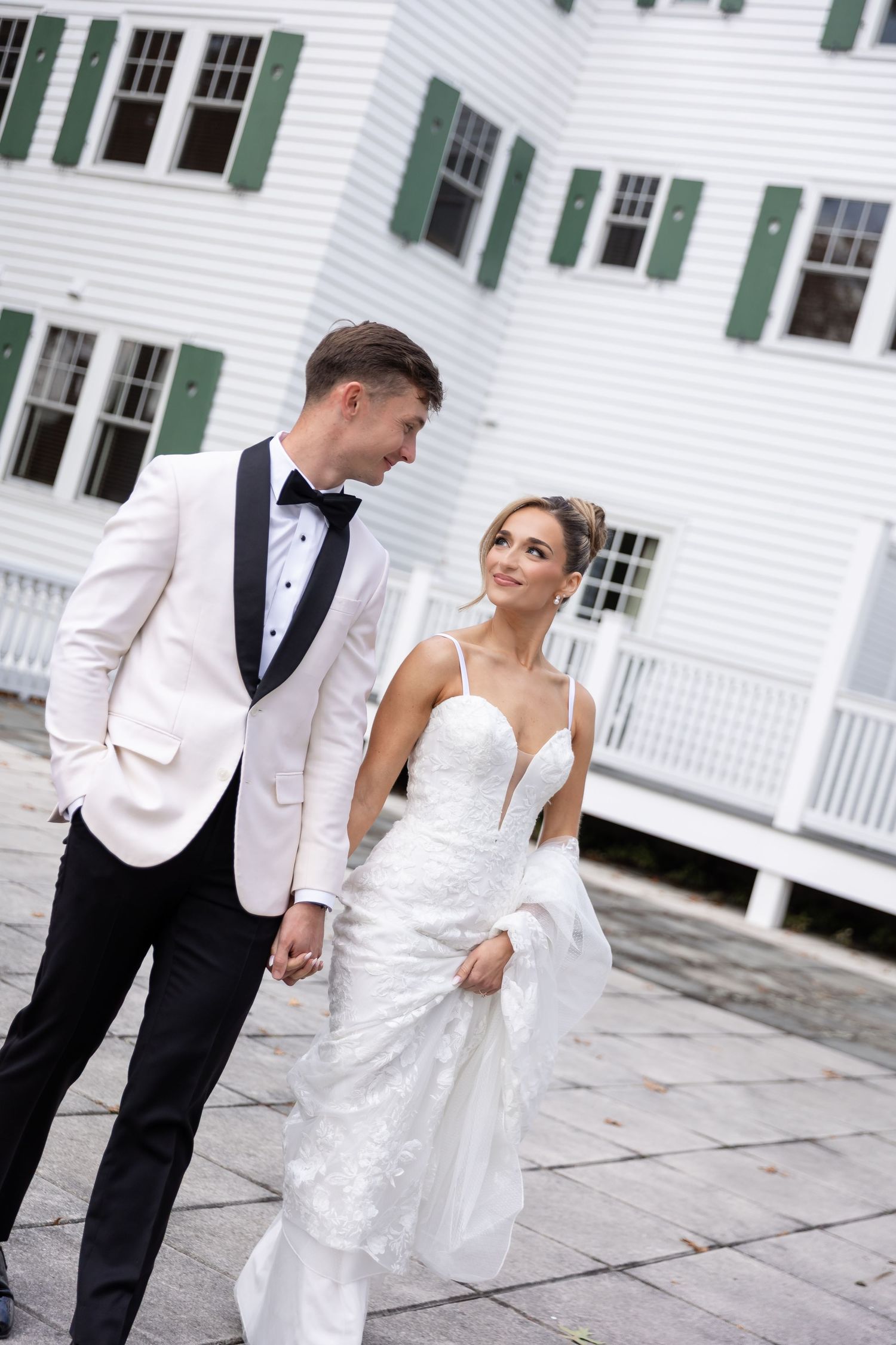 A couple in wedding attire poses together outside a classic white colonial building with green shutters.