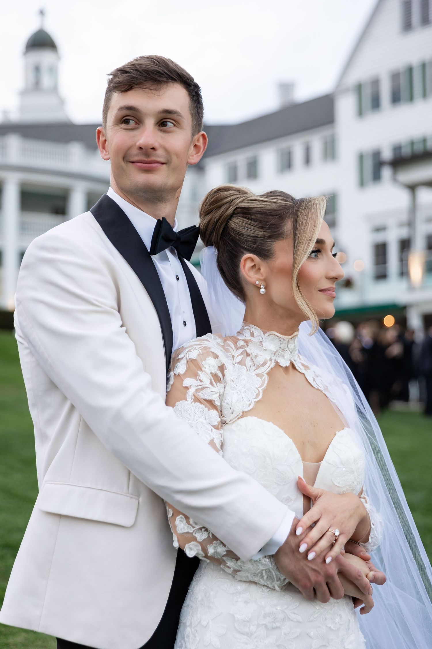 A romantic wedding couple embraces outdoors in front of a white building, with the groom wearing a white tuxedo jacket.