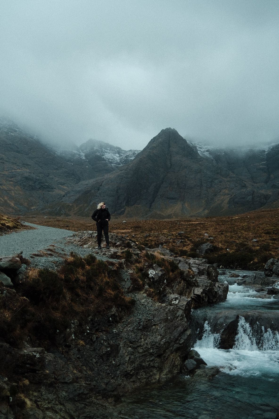 A solitary adventurer stands on rocky cliffs above rushing waterfalls with foggy mountains in the distance.