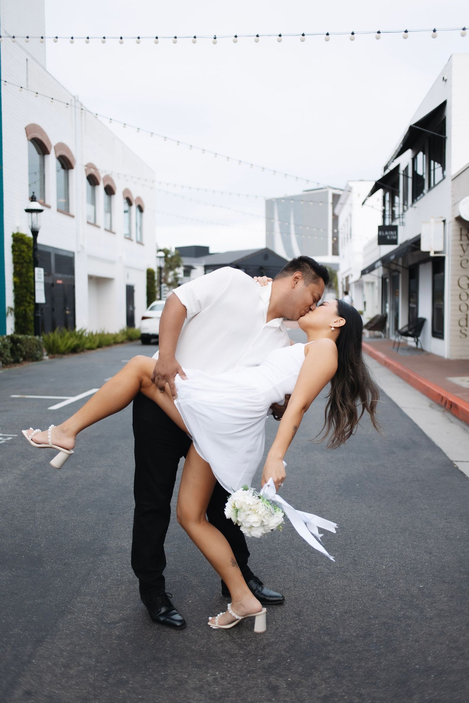 A romantic dip pose on a street lined with string lights and white buildings.