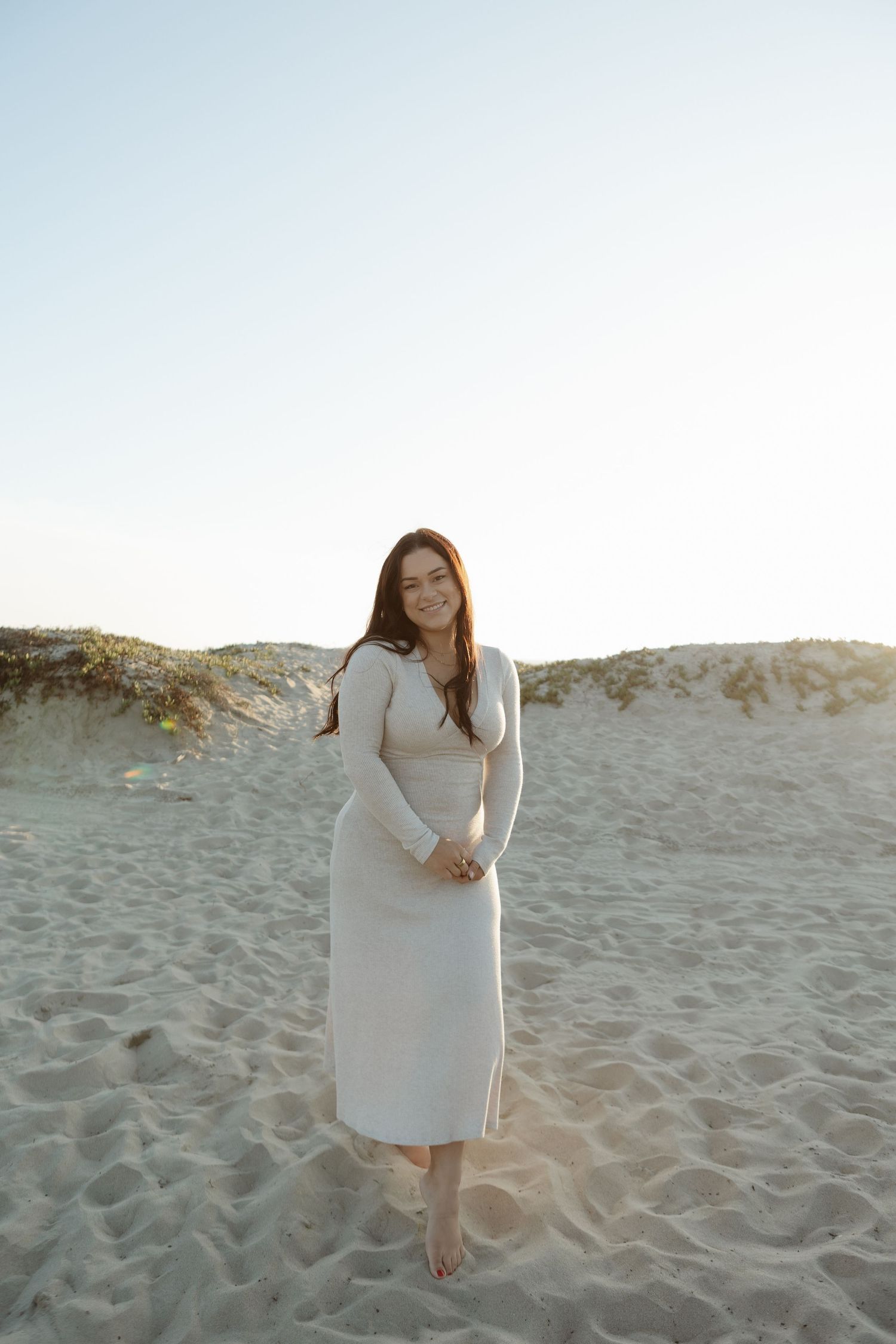 A person in a long white dress stands barefoot on a sandy beach at sunset.