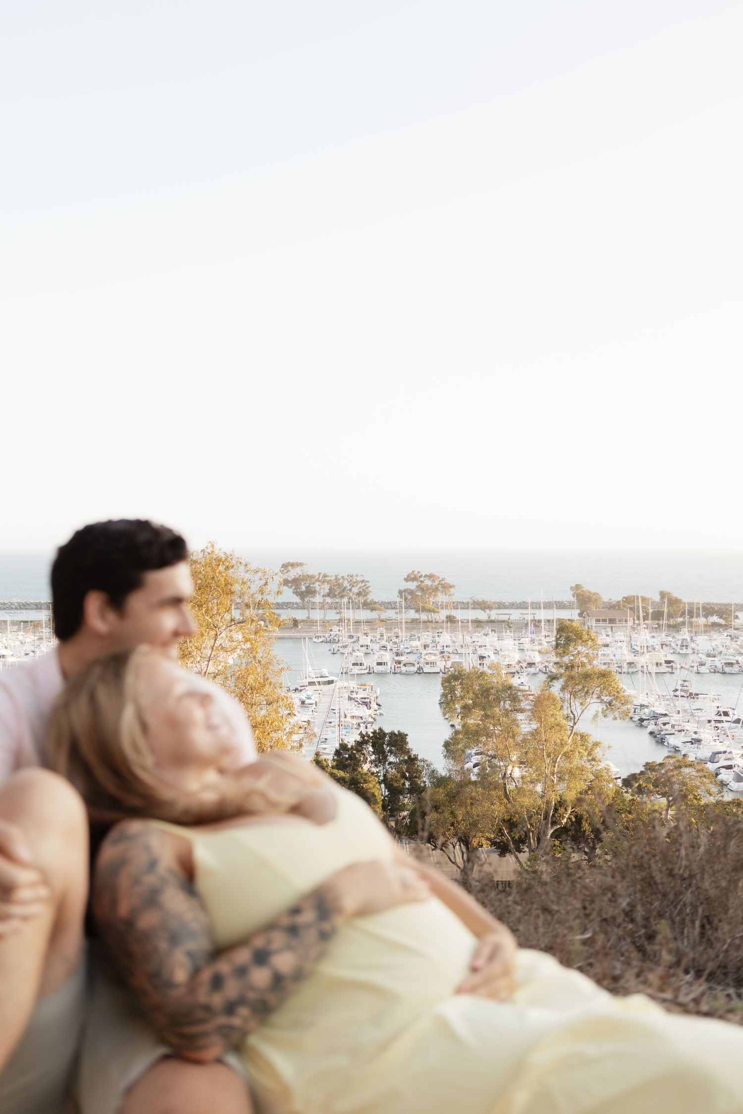 A couple shares an intimate moment overlooking a marina with boats and autumn trees in the background.