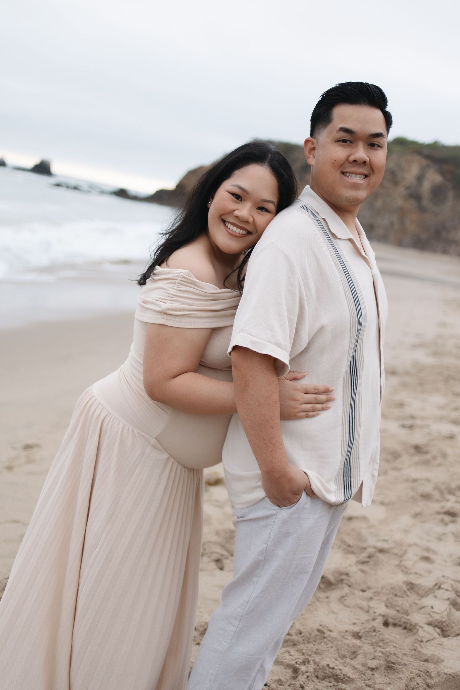 A couple poses together on a beach in light colored formal attire.