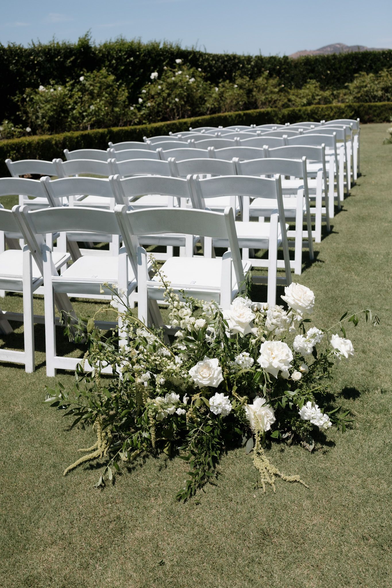 White chairs arranged in rows on a grassy lawn with floral decorations lining the aisle for an outdoor wedding ceremony.