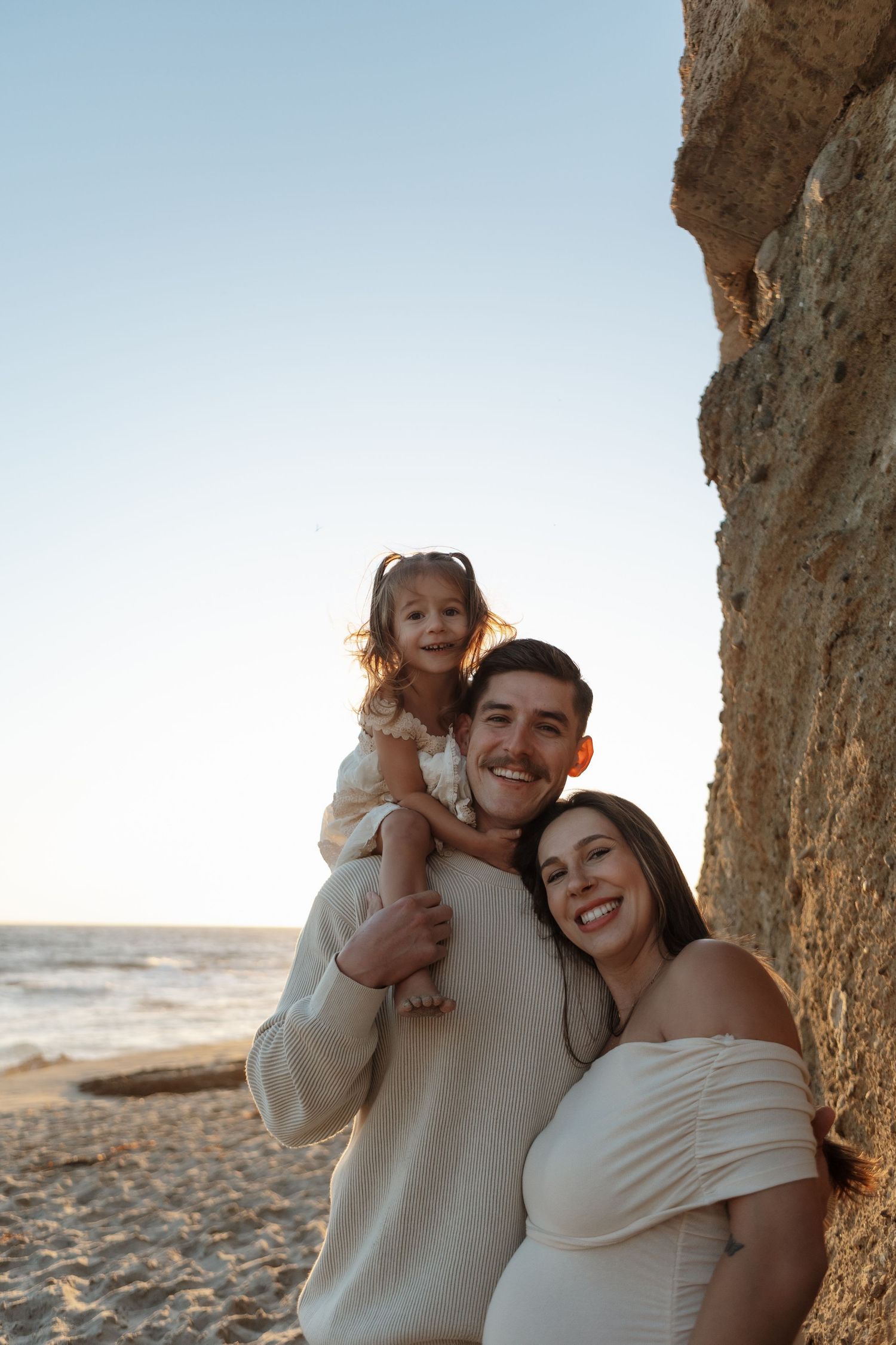 Family enjoying a tender moment together on a sandy beach at sunset with rocky cliffs in the background.