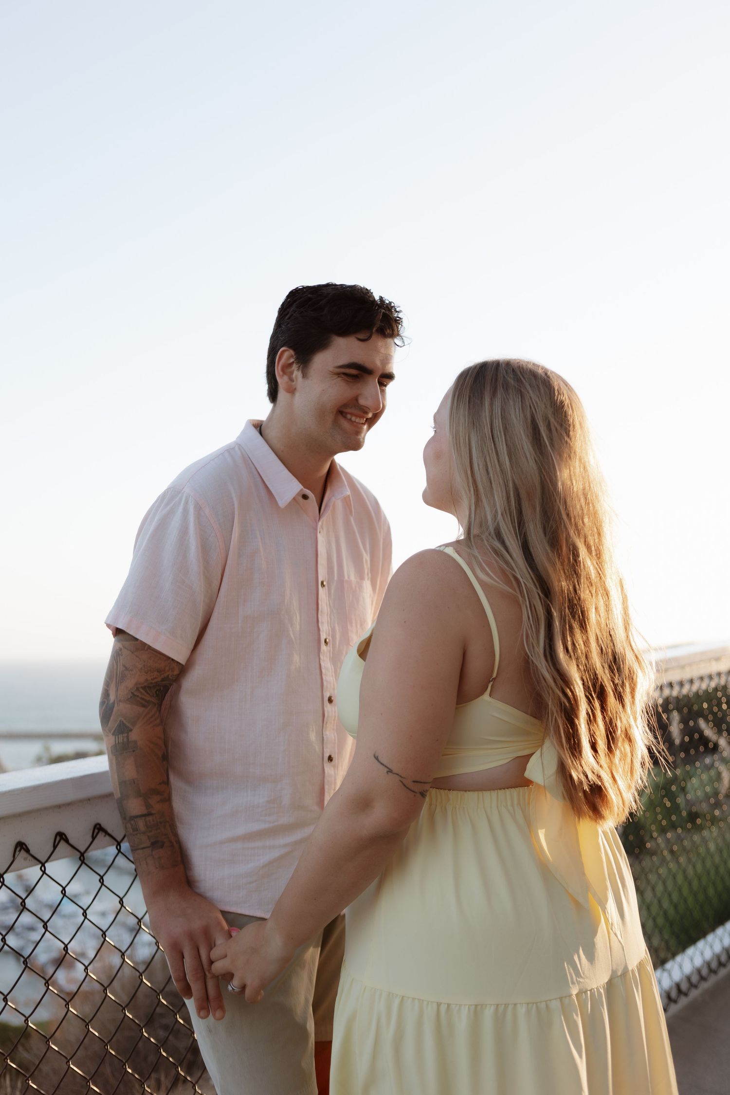 A couple in light summer outfits share a tender moment at sunset by a chain link fence overlooking a scenic view.