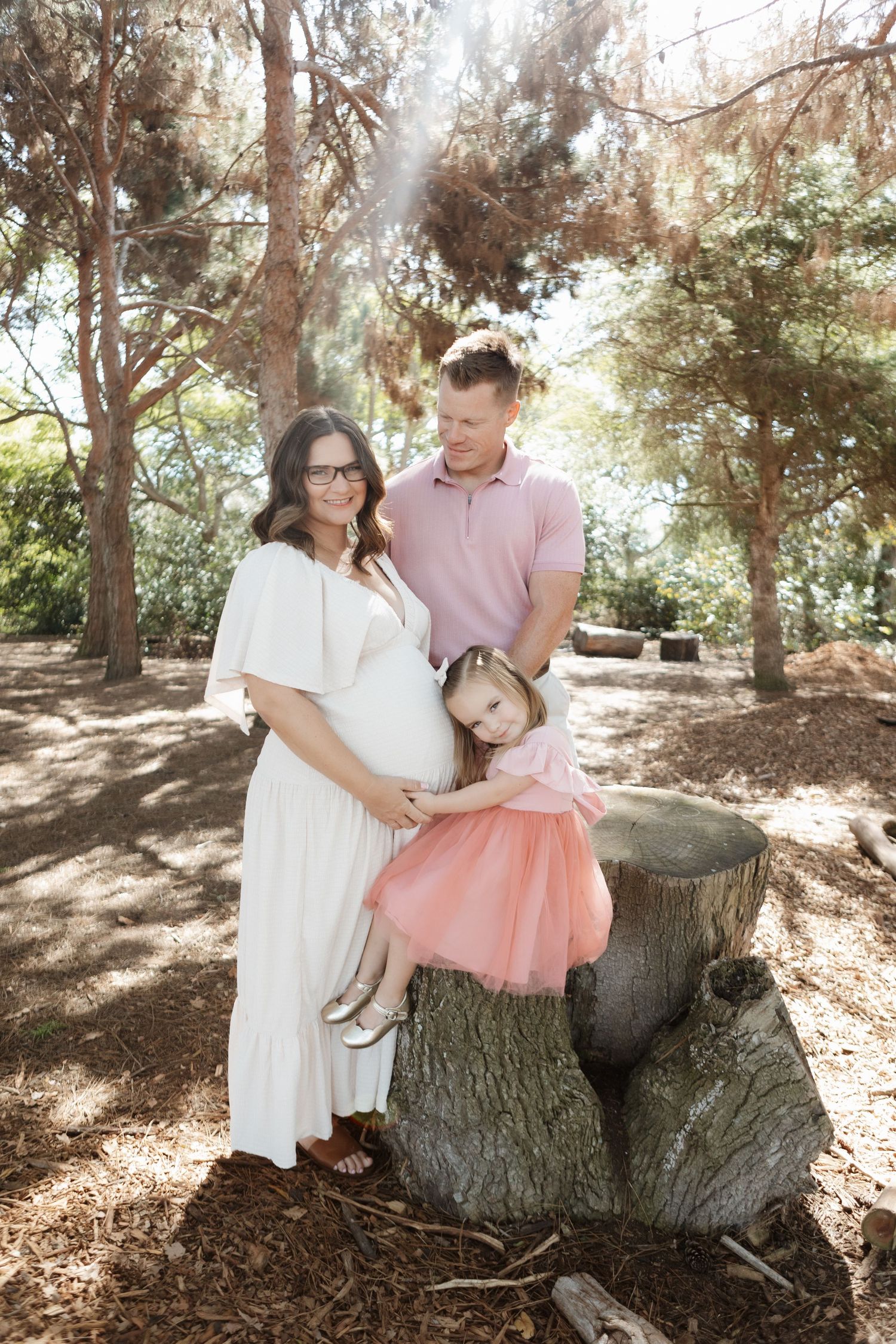 A family poses together in a dreamy forest setting with sunlight filtering through the trees.