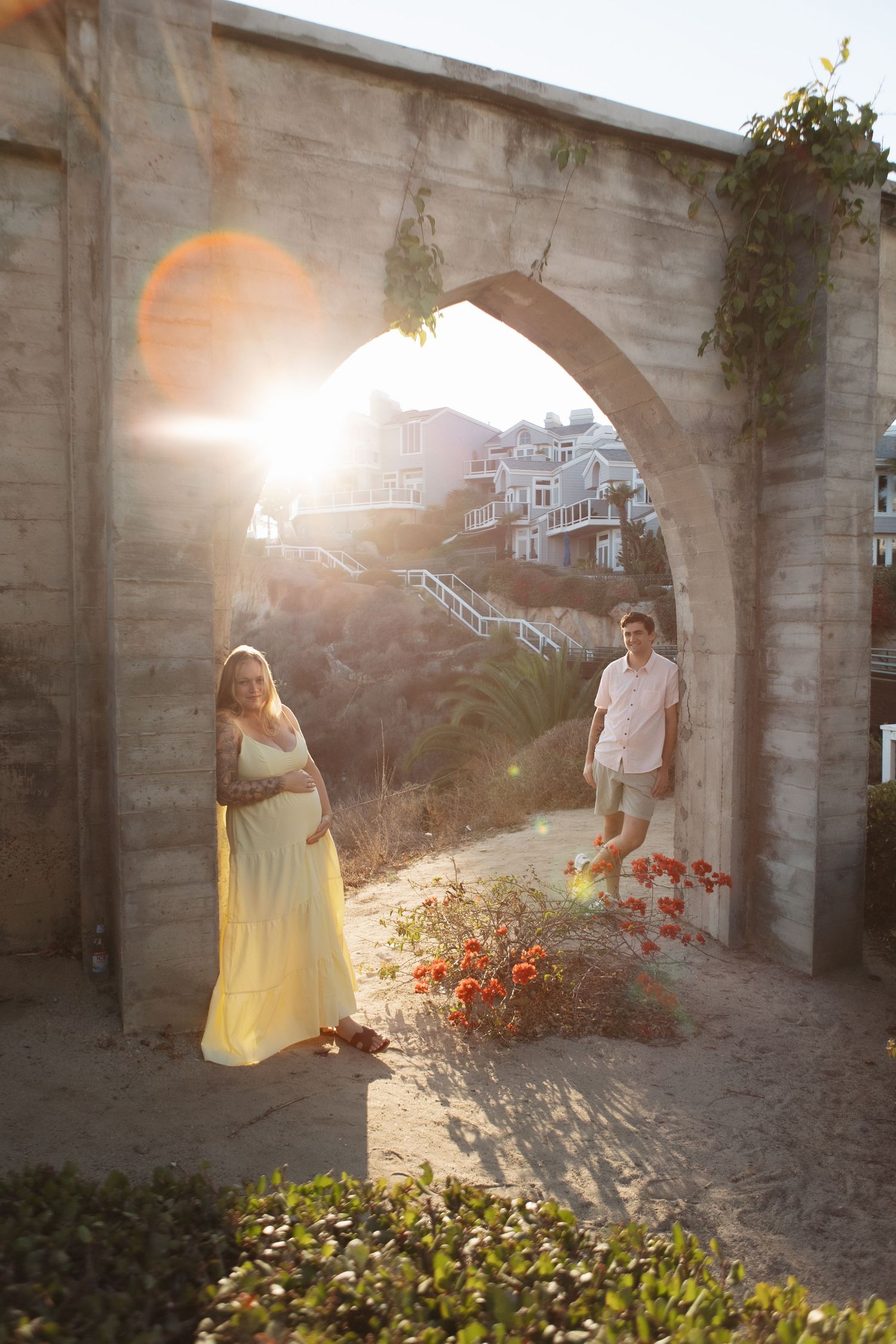 Couple stands in stone archway at sunset with red flowers and coastal homes in background.
