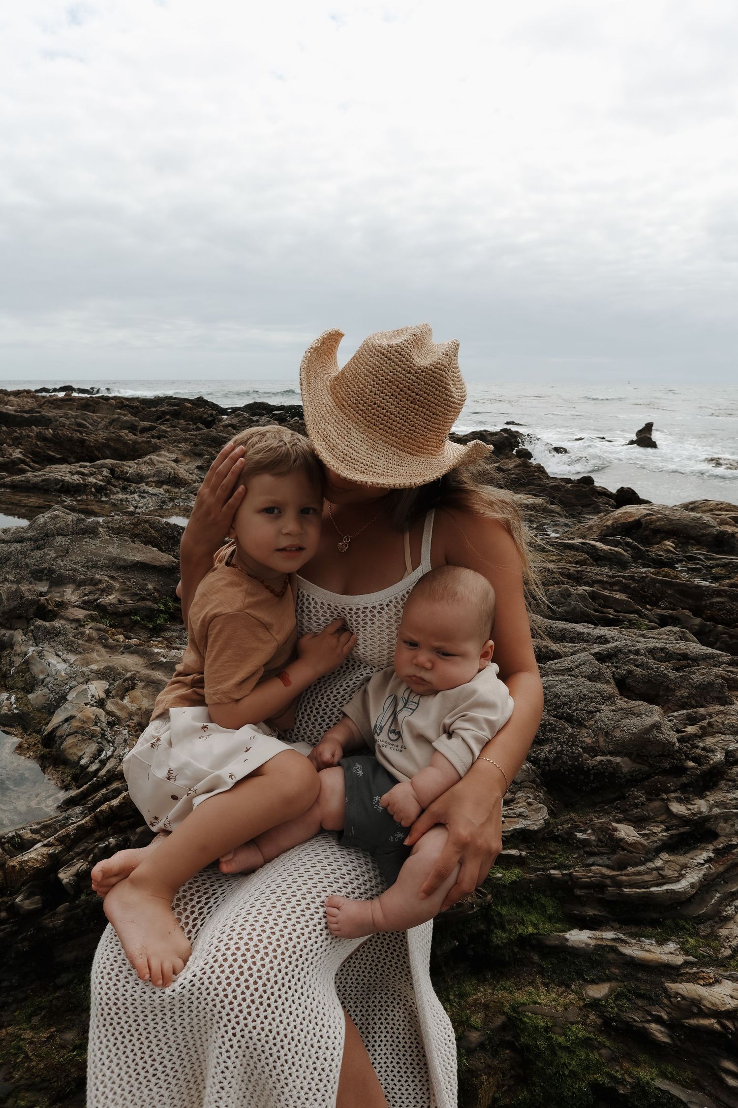 A tender family moment captured on rocky coastal cliffs with ocean waves in the background.