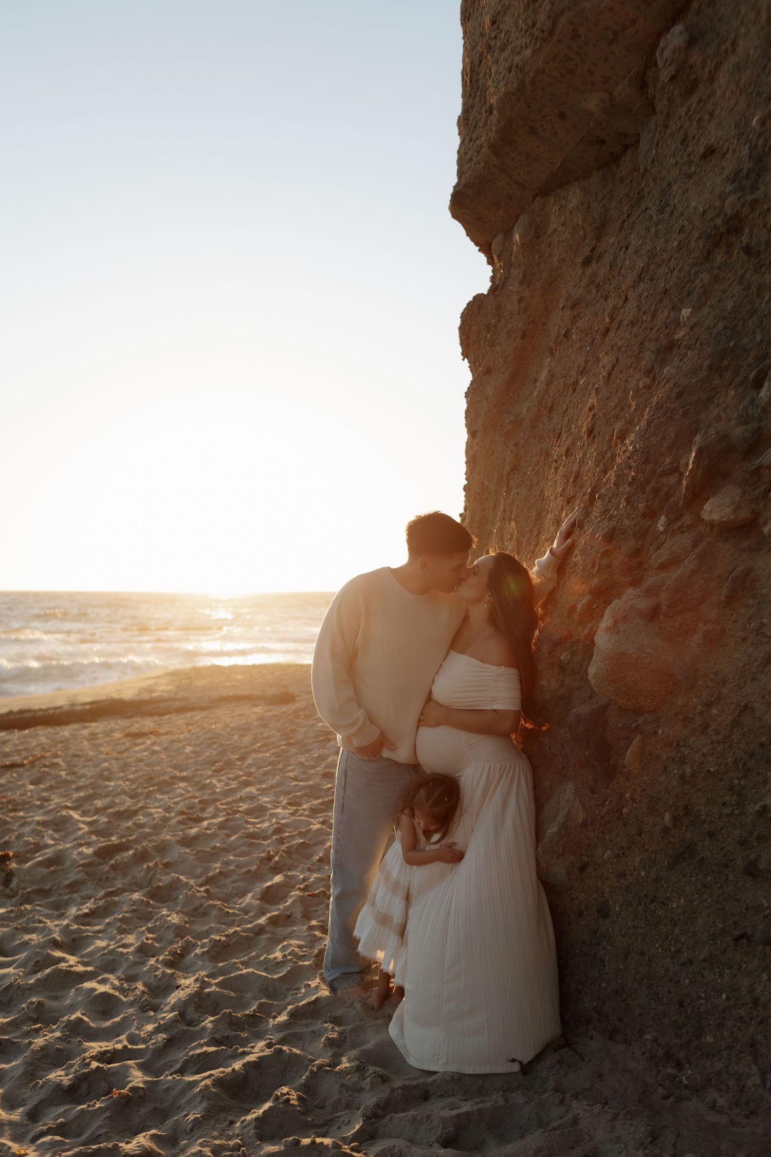 A couple embraces on a sandy beach at sunset, silhouetted against a rocky cliff backdrop.