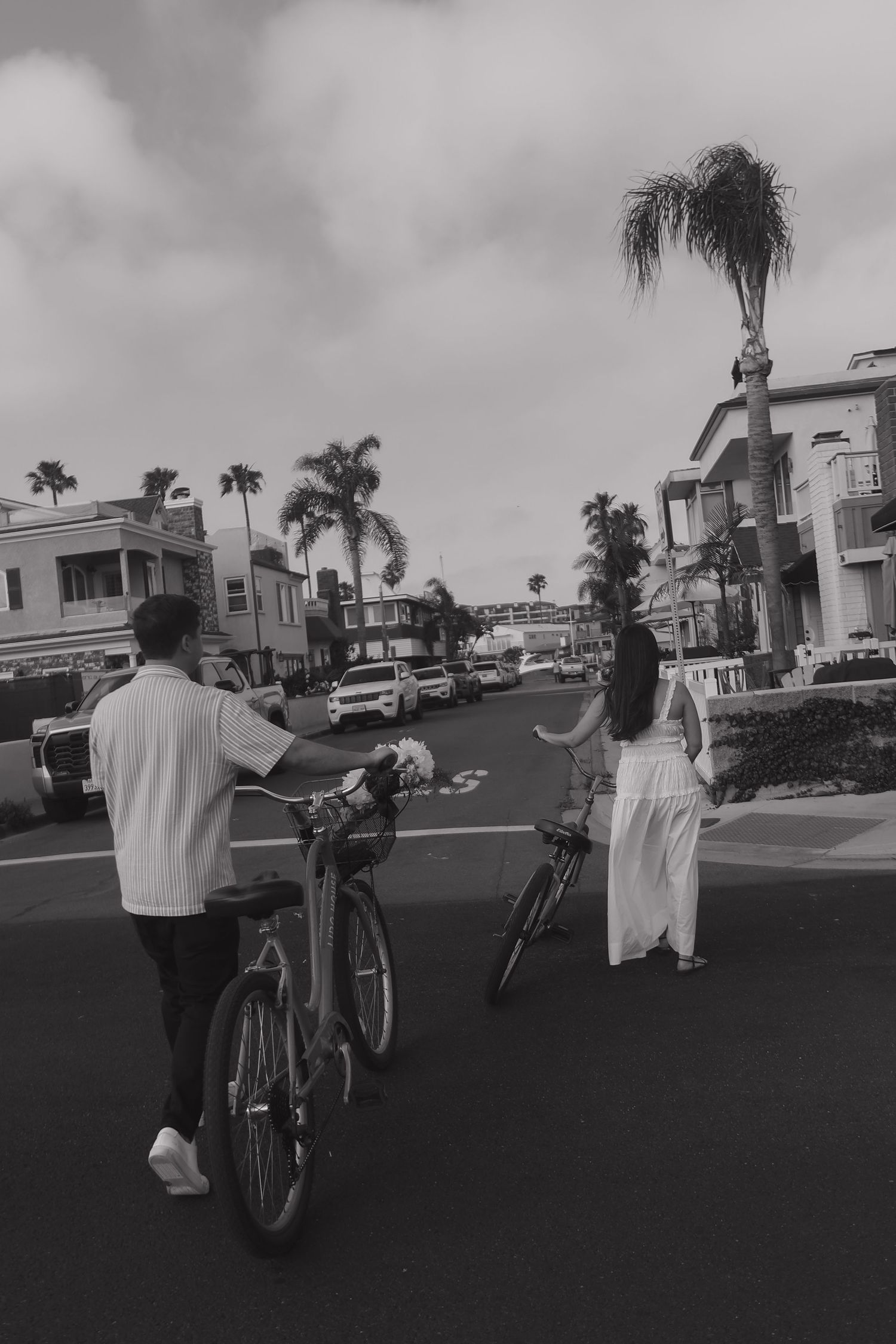 Black and white photo of people cycling on a suburban street with houses and palm trees visible.