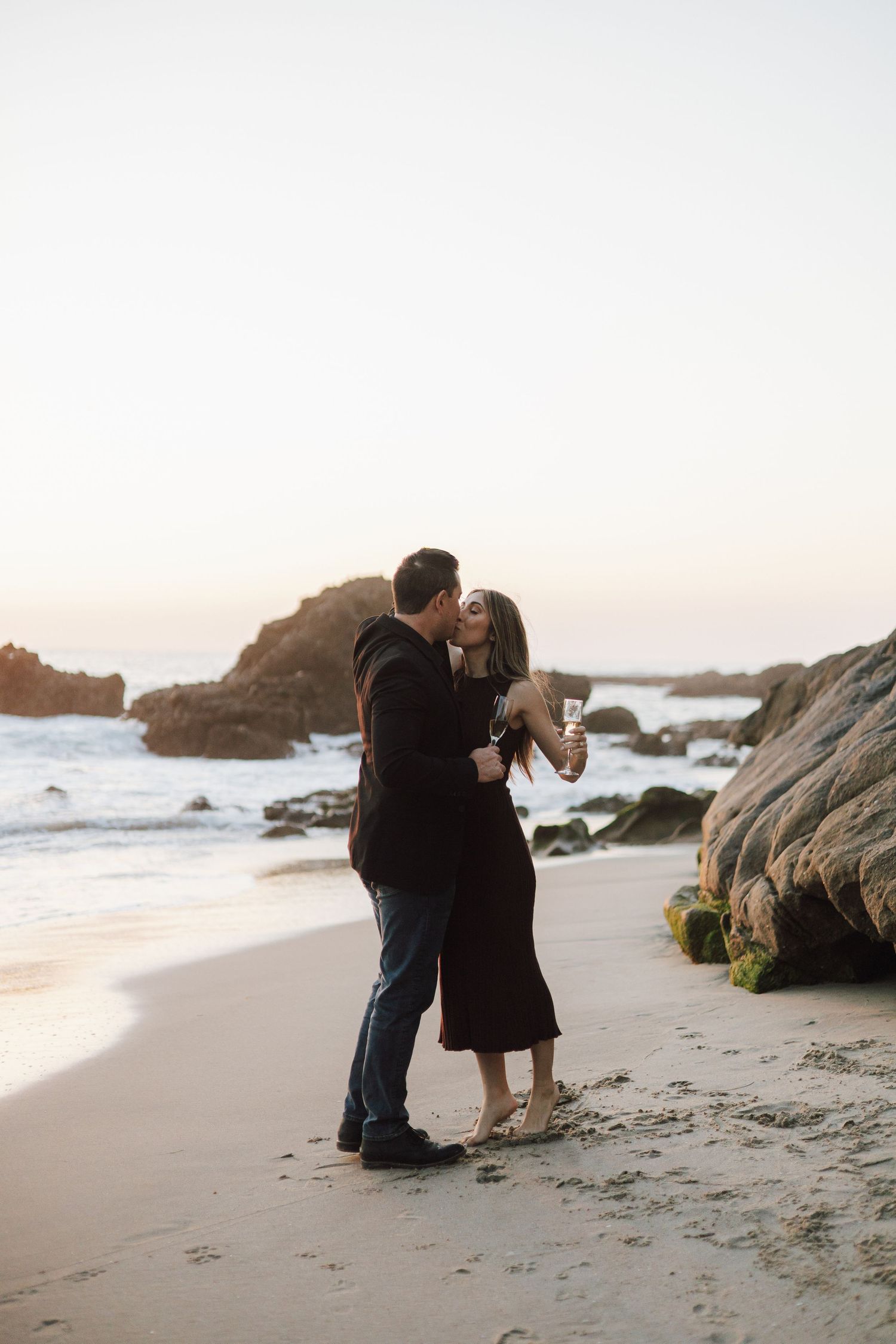 A couple embraces on a rocky beach at sunset with waves crashing against coastal rocks in the background.
